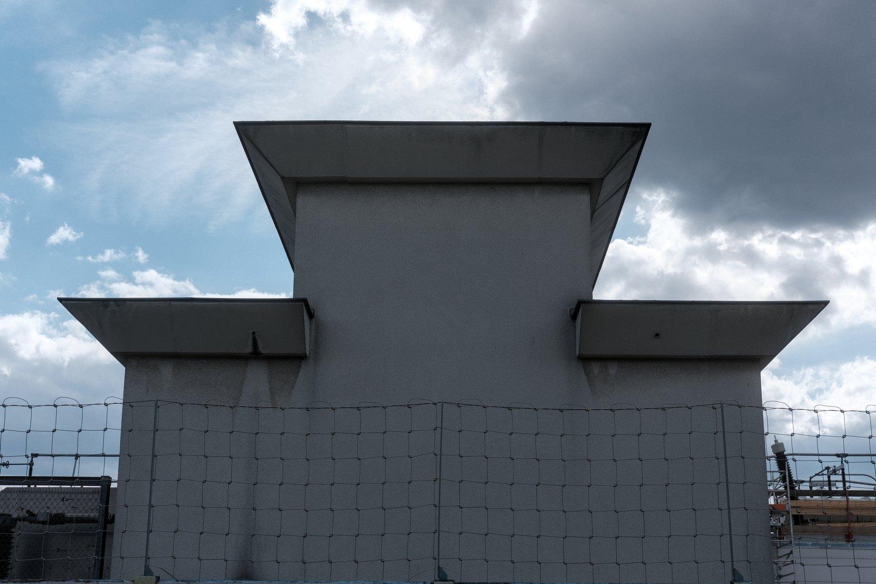 Photo horizontale. Un bâtiment derrière un grillage de protection. Il a la forme d’un podium avec les marches des côtés de la même hauteur. Au dessus, un ciel bleu à gauche et un gros nuage gris à droite
