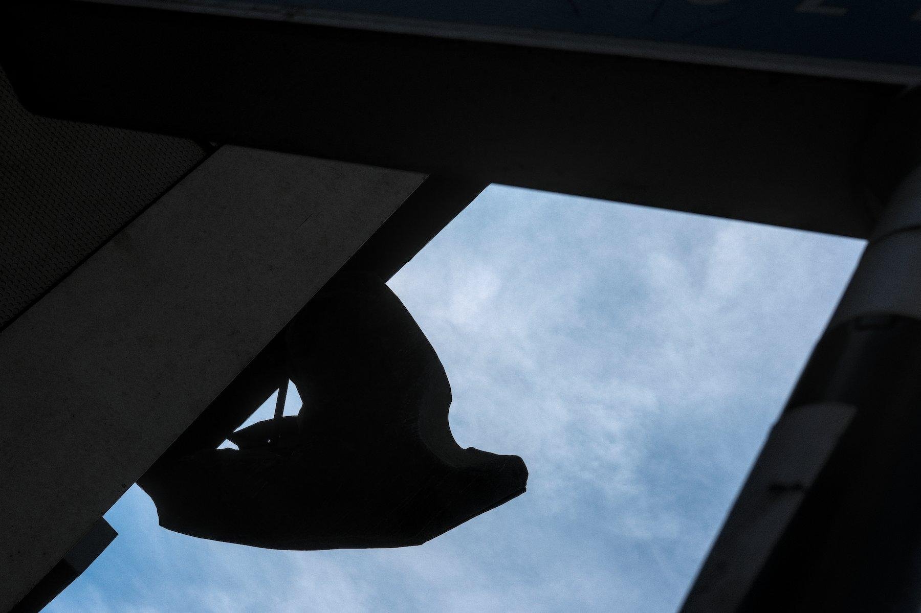 Photo horizontale. Encadré par des structures en contre jour, une sculpture de colombe accrochée à un mur se dessine en contre-jour sur un ciel bleu et blanc