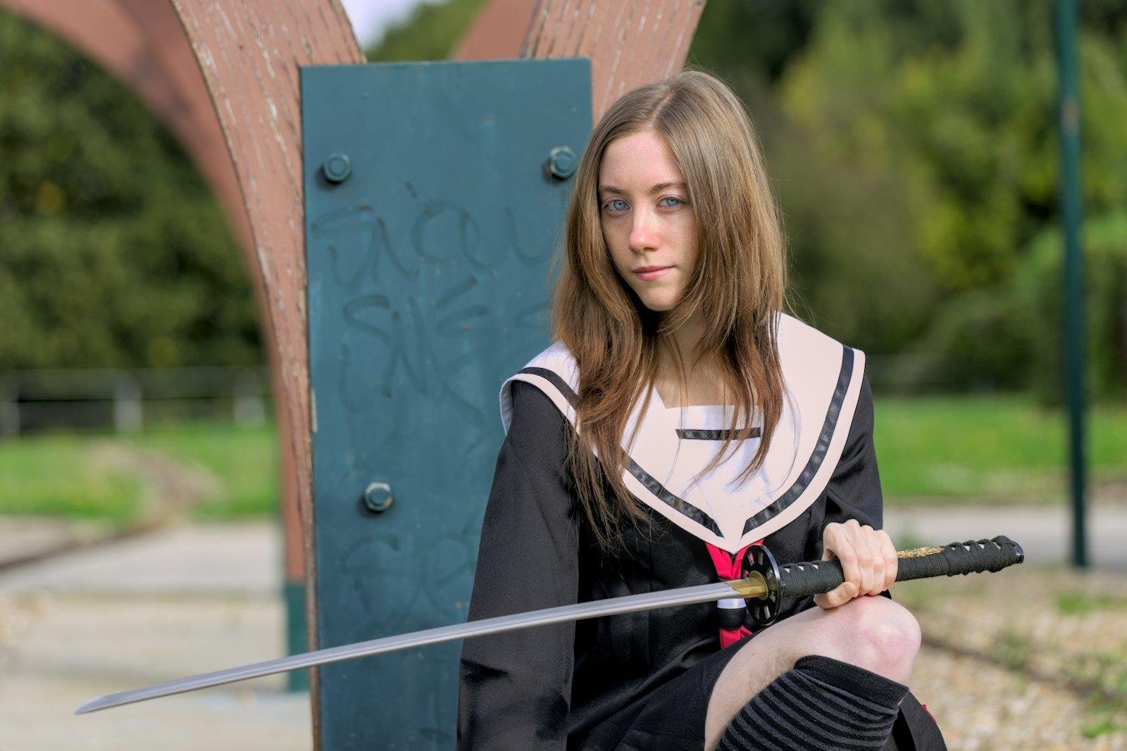 Une jeune femme en tenue d’écolière, accroupie, tenant un katana de la main gauche, et dont la lame repose sur sa cuisse droite, regardant la caméra d’un air décidé