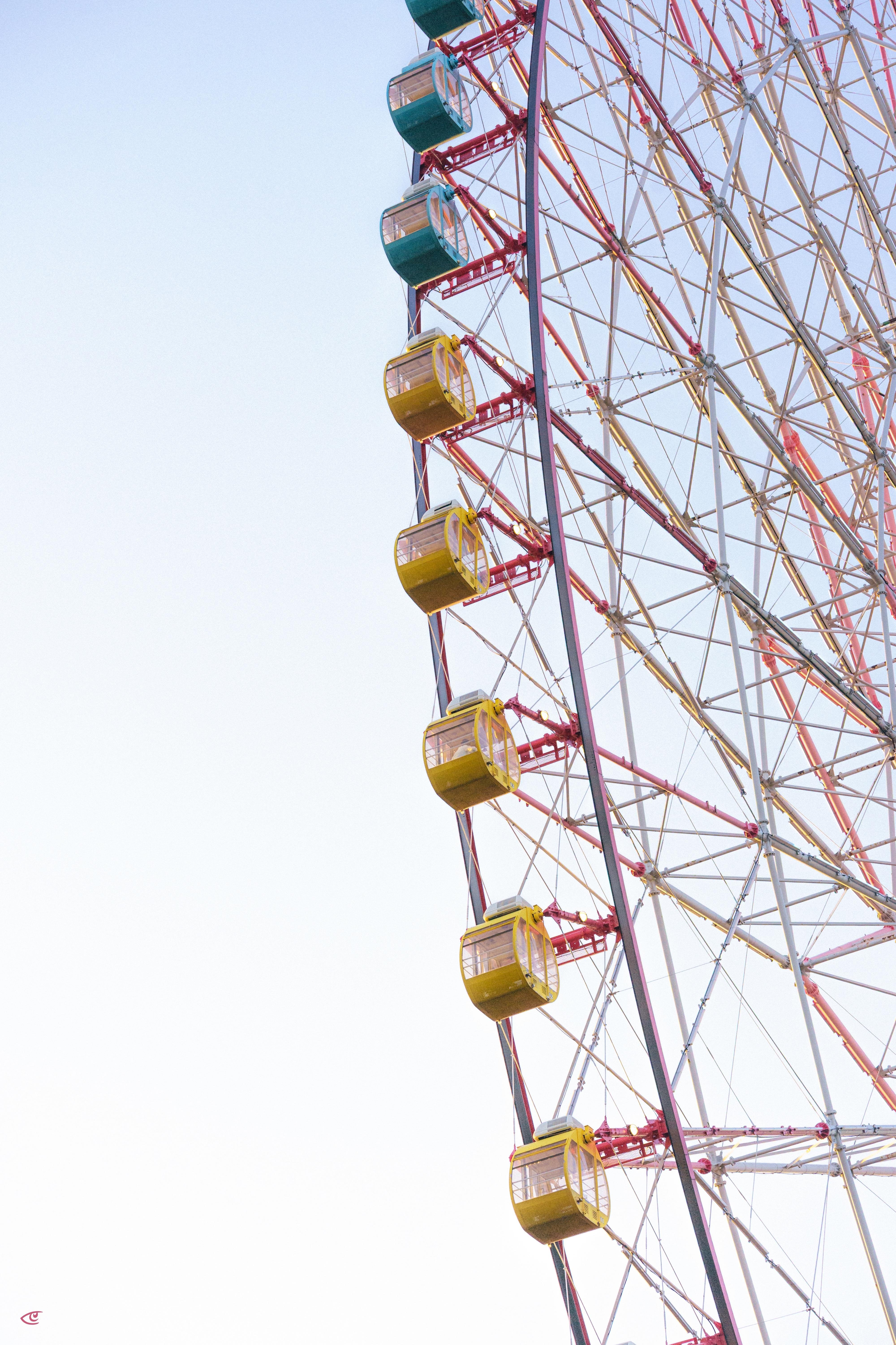 Right side of a large Ferris wheel with red metal framework and several gondola cabins in yellow and teal, set against a pale sky with open white space on the left.