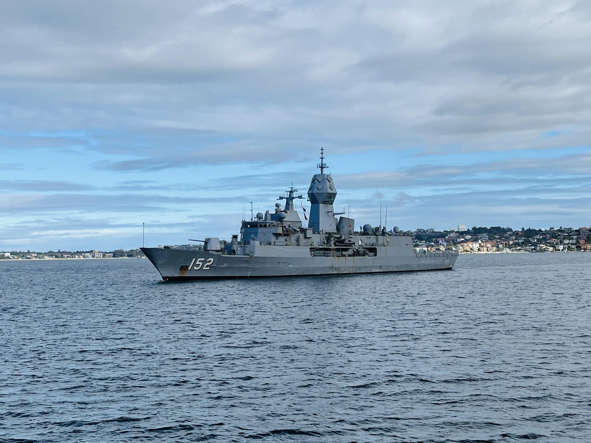 Gray naval ship with the number 152 on its bow, sailing in calm waters. Background features a coastal cityscape with low-rise buildings and rolling hills under a cloudy sky.