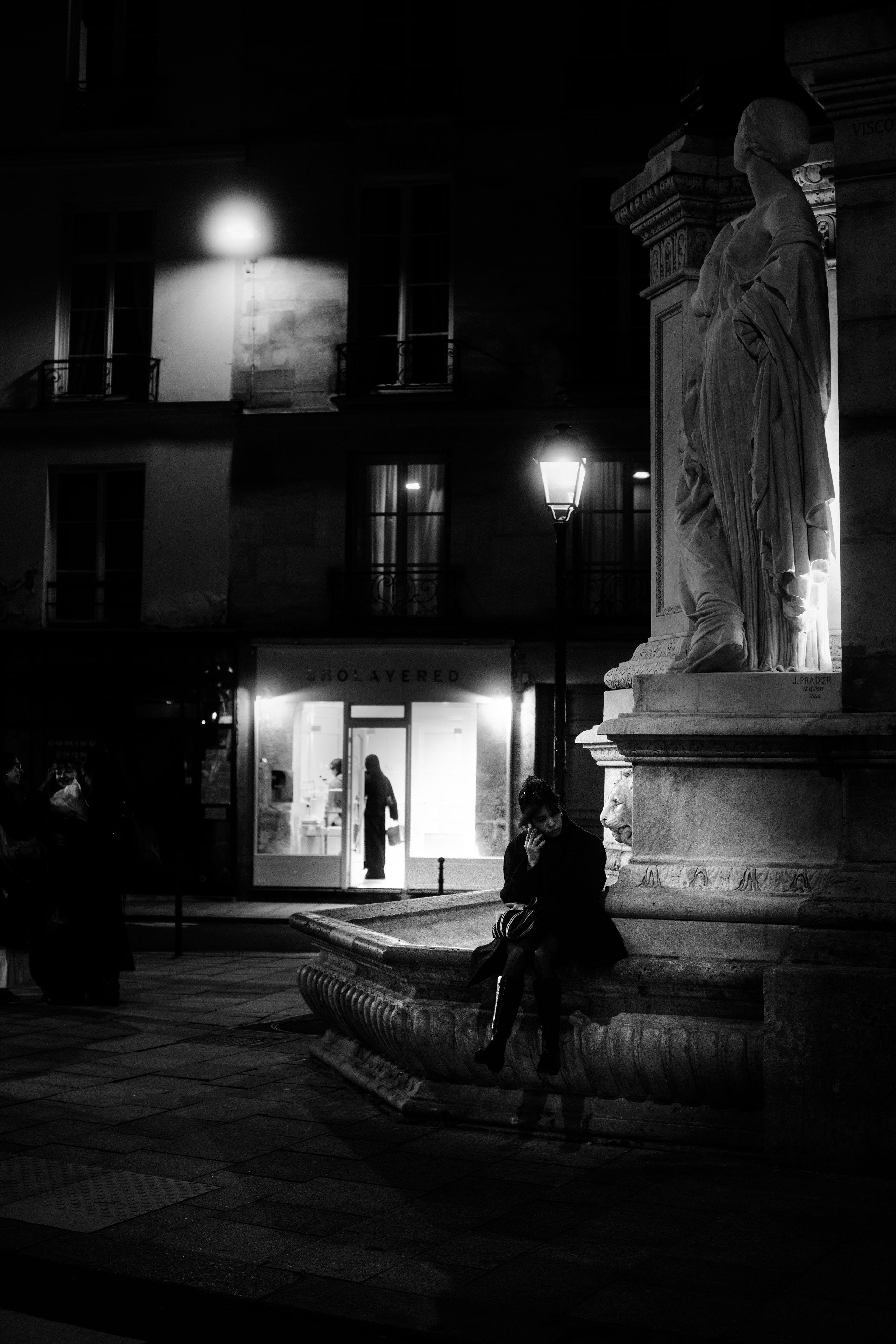 Photo verticale en noir et blanc, de nuit. Une jeune femme est assise sur le rebord d’une fontaine surmontée d’une statue. Au fond, de la lumière émanant d’une vitrine d’un commerce avec la silhouette d’une femme dans l’encadrement de la porte d’entrée. Autour de ce commerce, un immeuble