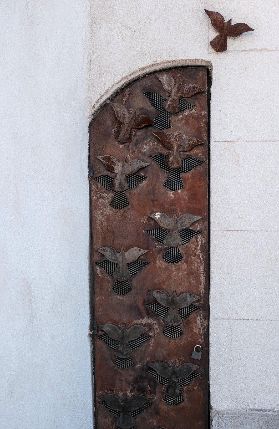 Photo verticale. Une porte en bois qui semble coupée en deux sur la longueur. La surface est sculptée de silhouettes d’oiseau vu de dessus, ailes déployées. Un oiseau en bois vu de dessus ailes déployées est accroché juste dans le coin supérieur droit de la porte, là où le sommet aurait du être si la forme de la porte était complète