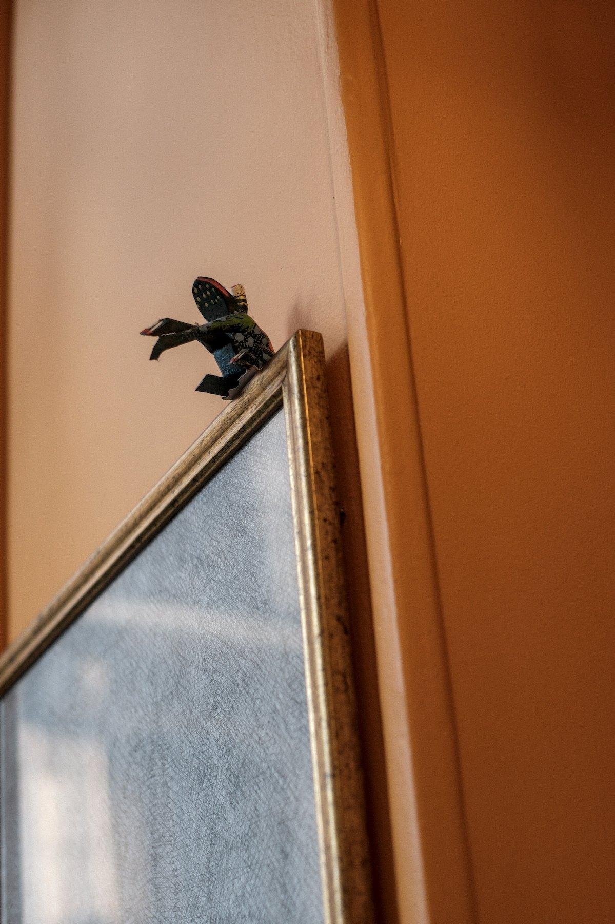 Photo verticale. Sur un mur orange, un tableau est fixé (on ne voit pas l’œuvre) et une petite figurine de crapaud est collée sur le bord du cadre