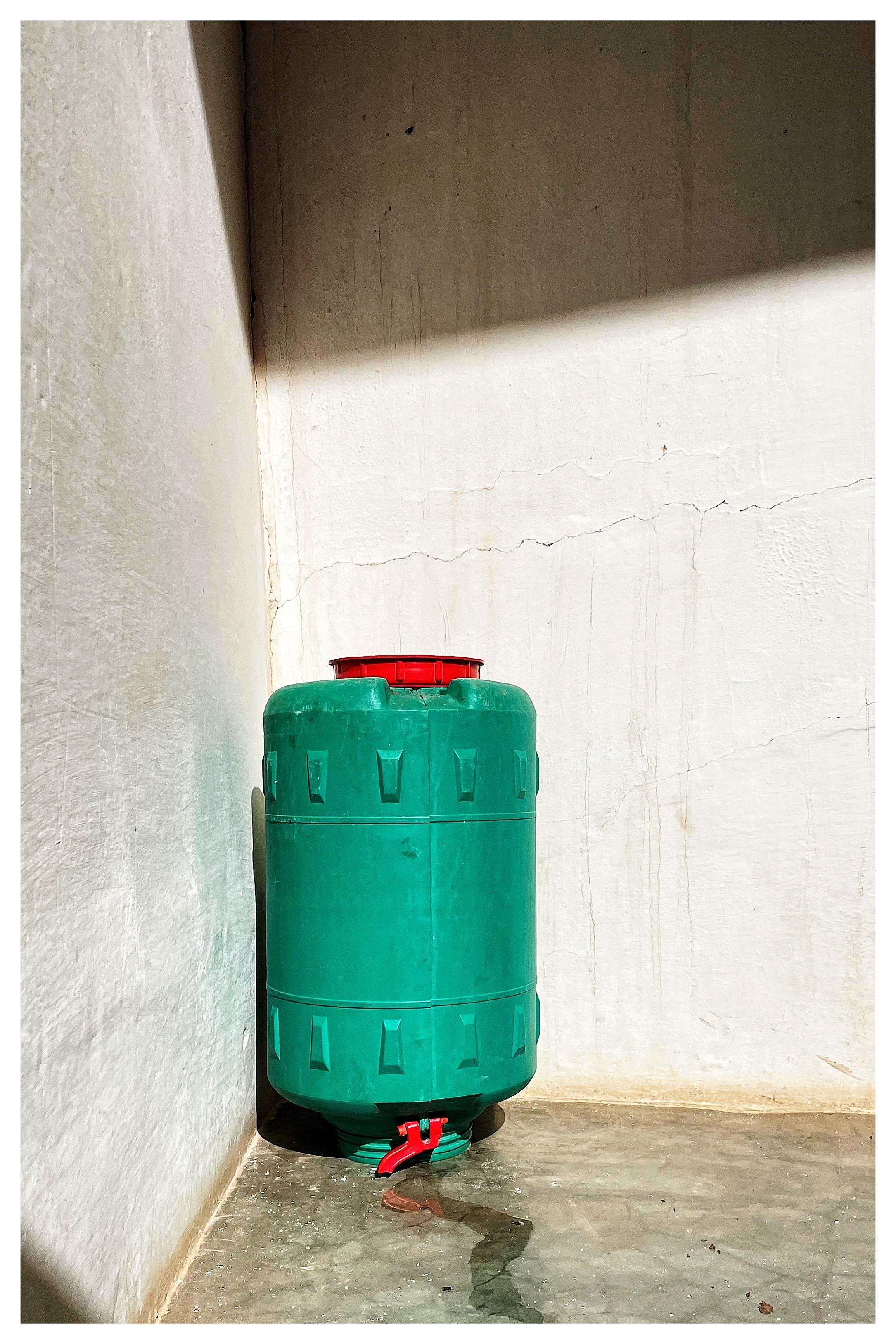 Large green plastic water tank with a red lid and red spout at the bottom, placed in the corner where two white walls meet; strong diagonal sunlight casts shadows on the walls and concrete floor.