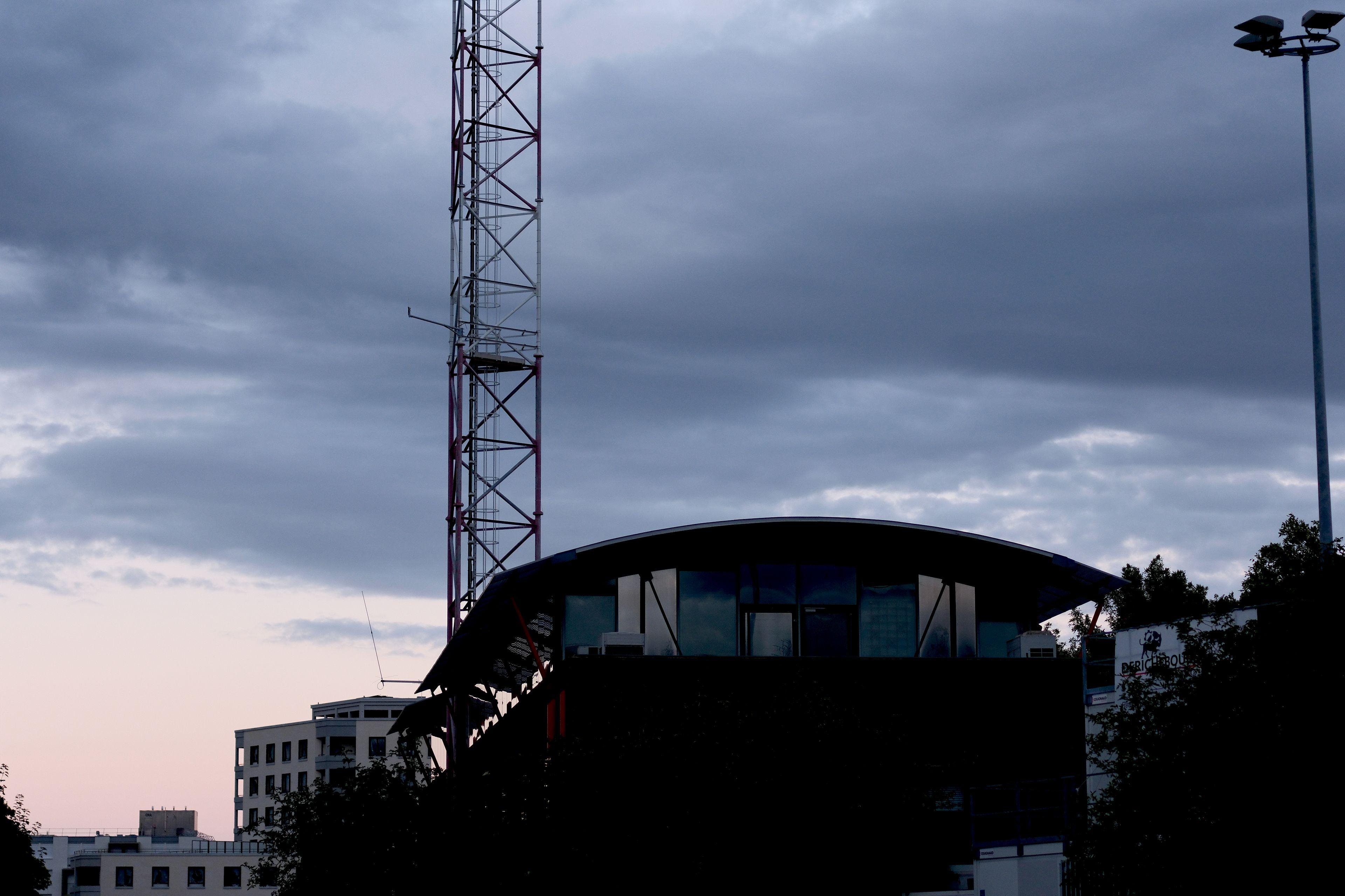 Photo horizontale. Un bâtiment dont le toit est en arc de cercle. Le début d’une tourelle faisant penser à une grue (mais c’est pour des antennes) sur fond de ciel de crépuscule