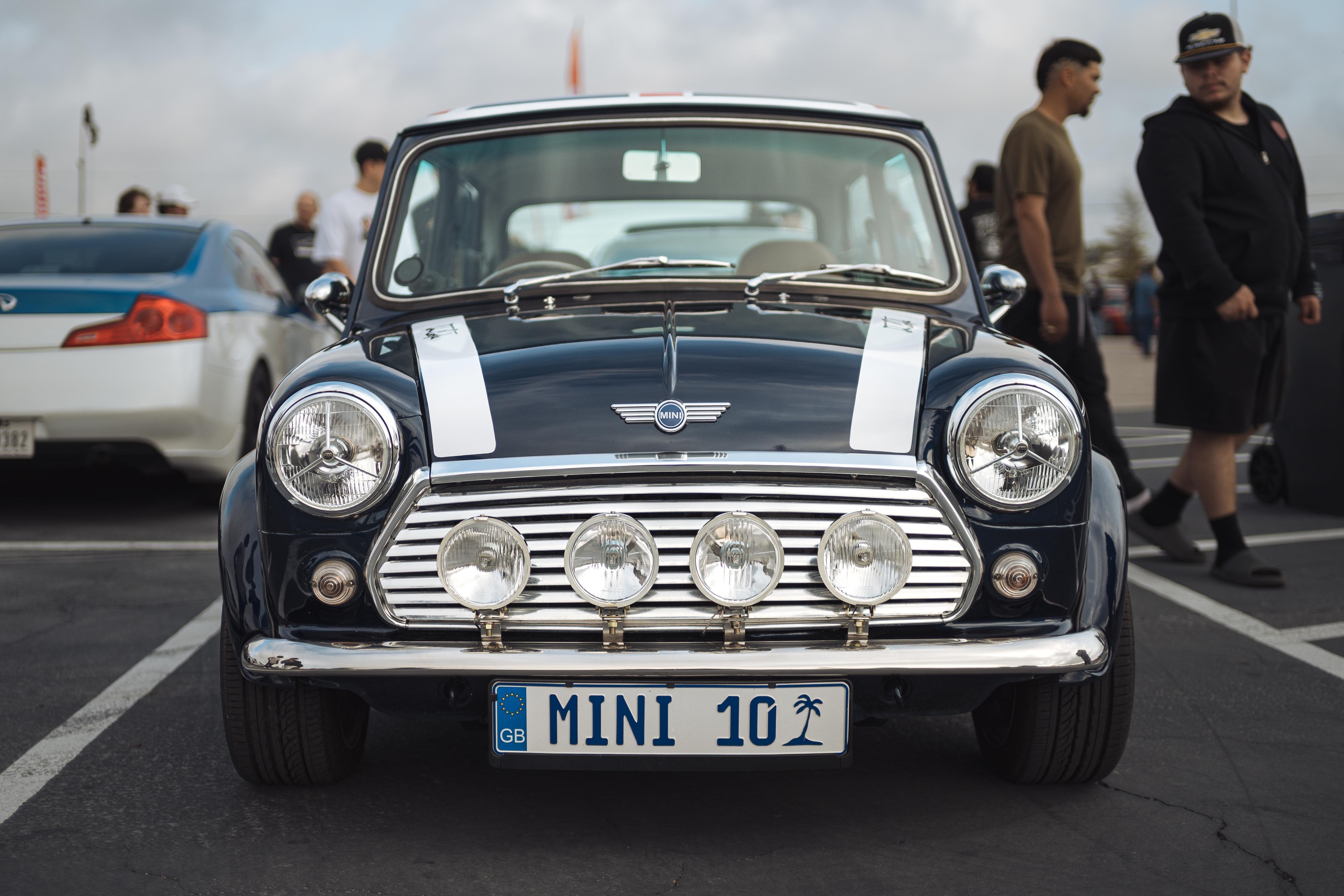 This image shows a classic Mini Cooper car with a dark blue color and white racing stripes on the hood. It has a set of four prominent rally lights on the front grille and a license plate that reads "MINI 10."