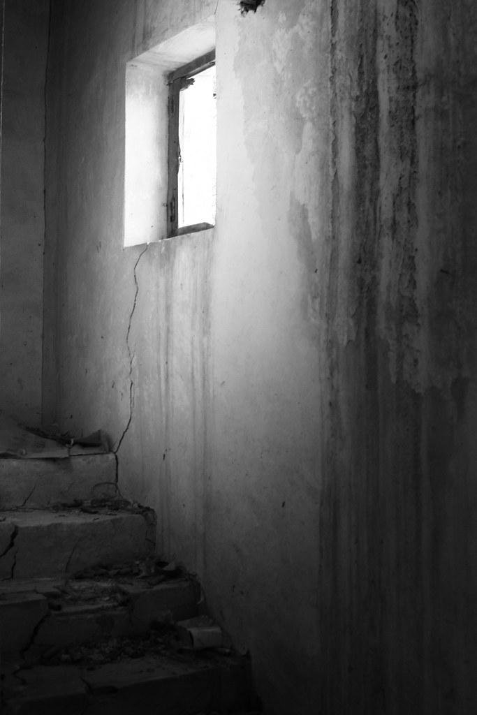 Una foto en blanco y negro de una pared de una casa en ruinas. La pared hace esquina y tiene unas escaleras en la esquina inferior derecha. En la pared hay una ventana sin cristales por la que entra la luz, que ilumina la pared manchada con un degradado suave