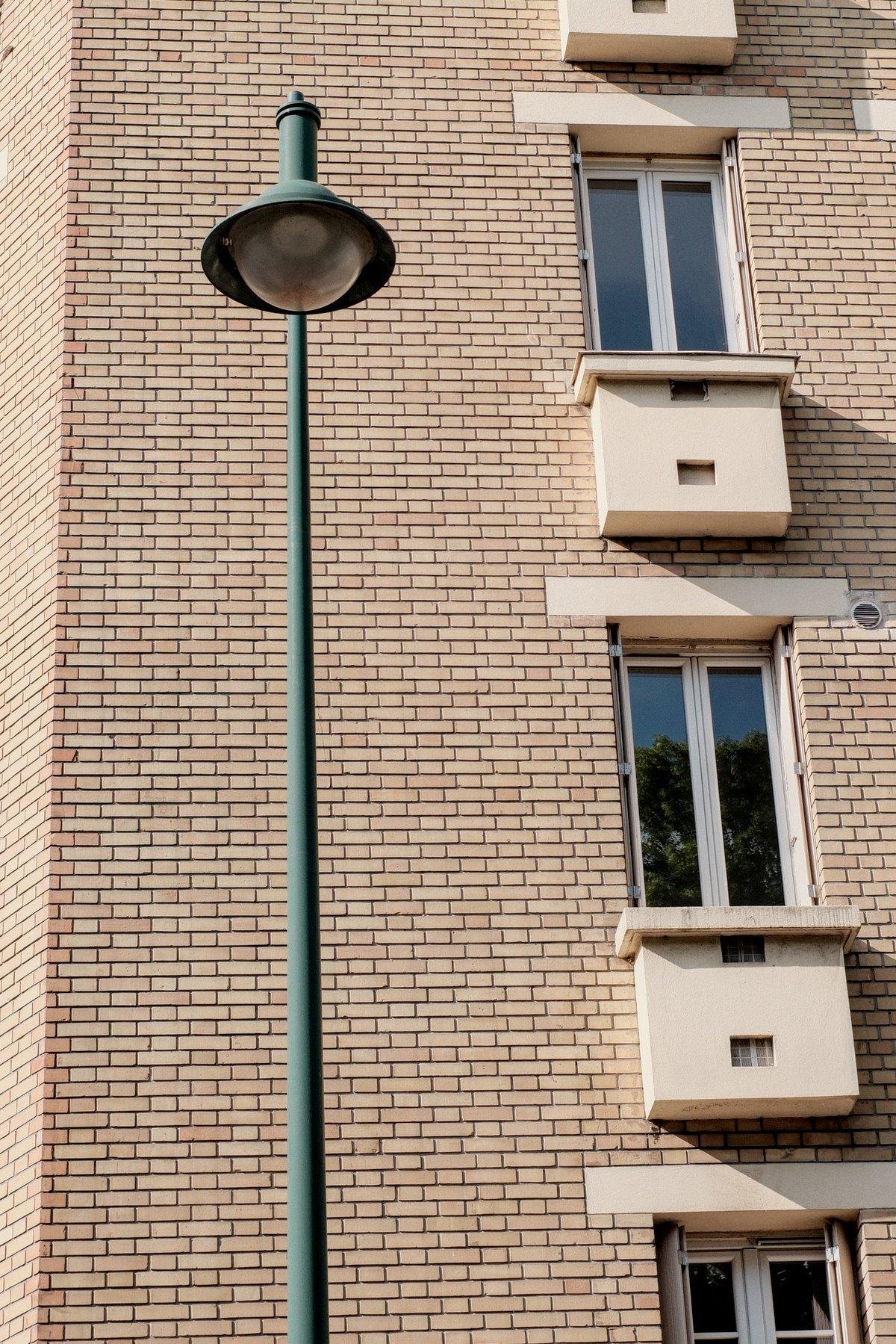 Photo verticale. Un bout d’immeuble en petite brique beige sombre, avec sur la droite, les une au dessus des autres, des fenêtres avec un garde manger, et sur la gauche, un lampadaire vert ressortant grâce au mur de briques derrière
