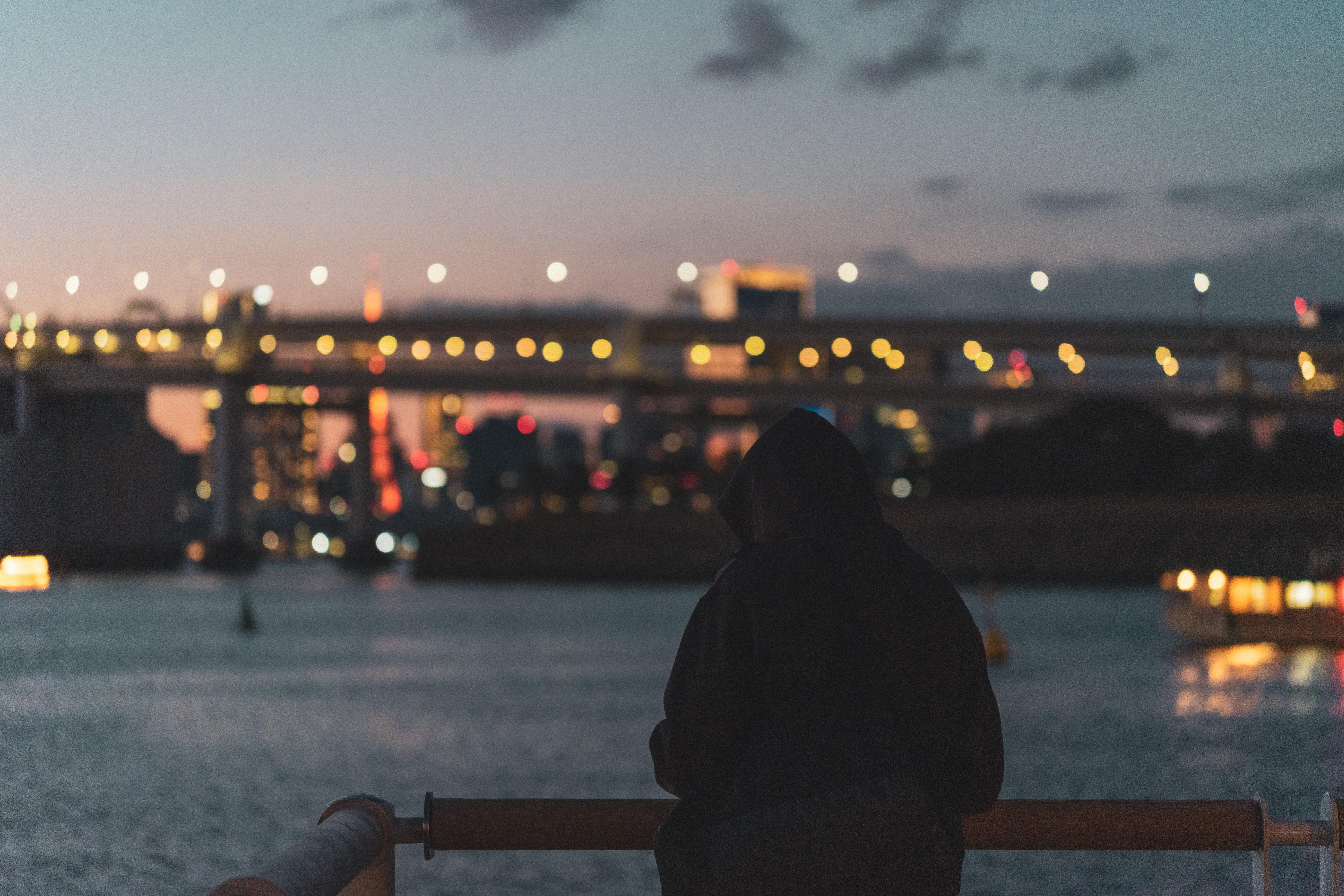 The image shows a person wearing a hooded jacket standing by a railing, overlooking a body of water. In the background, there is a bridge with lights, and some buildings with city lights are visible, suggesting it's taken during dusk or nighttime. The overall mood is serene and contemplative.