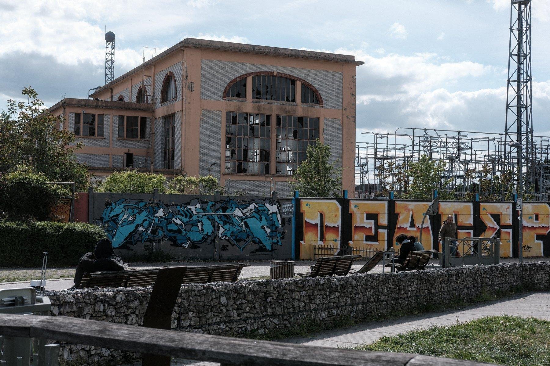 Photo horizontale. Un bâtiment gris et orange pâle, avec de grande baie vitrées mais dont plein de carreaux sont cassés, sur la droite, un pylône électrique. Devant, un grand mur de protection en béton, plein de gros graffitis. Encore devant, des gens assis sur des banc publiques devant un muret fait en pierre tenues dans une cage de fer