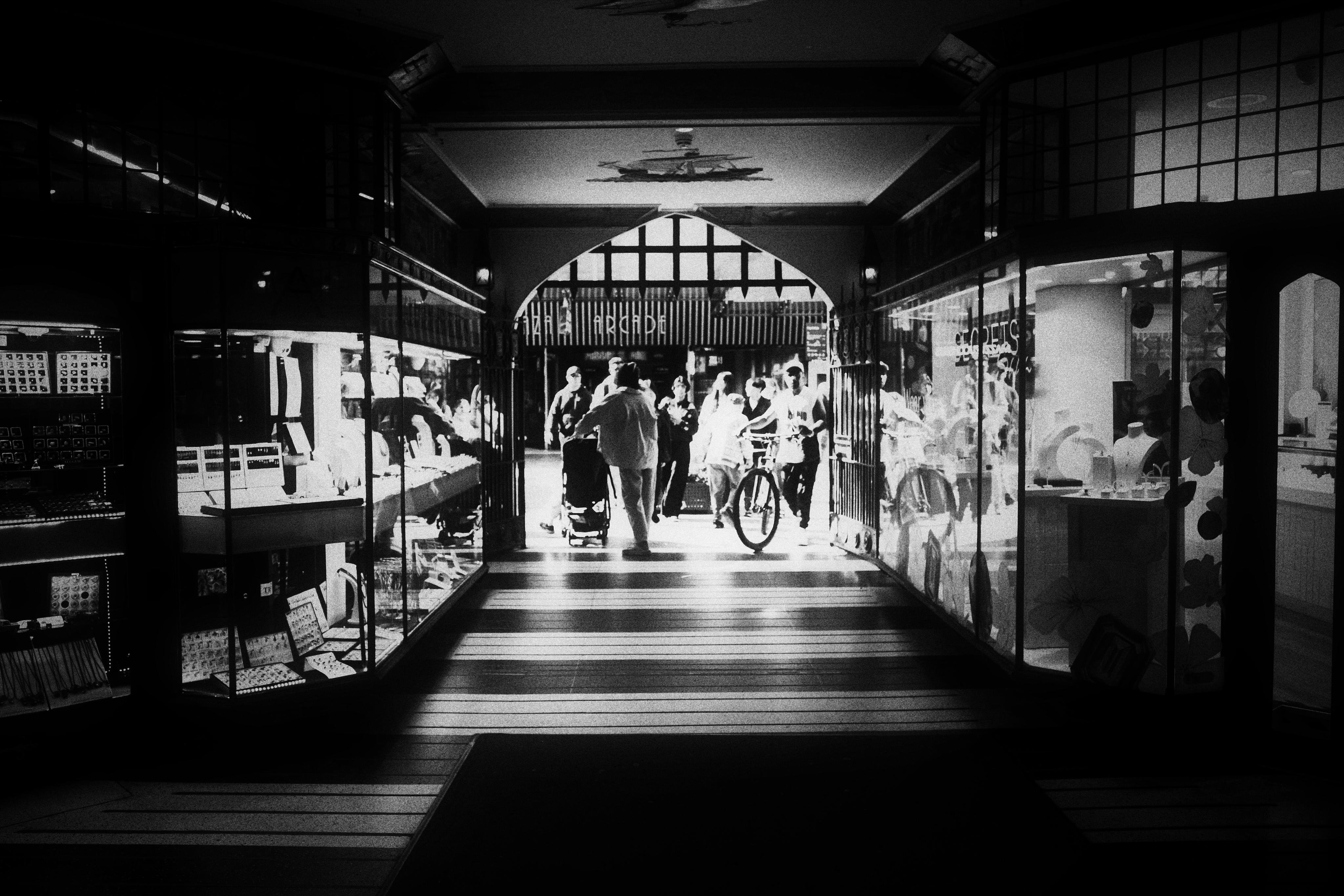 Black-and-white view down a covered shopping arcade with glass display windows on both sides; several people walk toward a bright exit at the far end, including an adult pushing a stroller and a person holding a bicycle, with striped light and shadow across the floor.