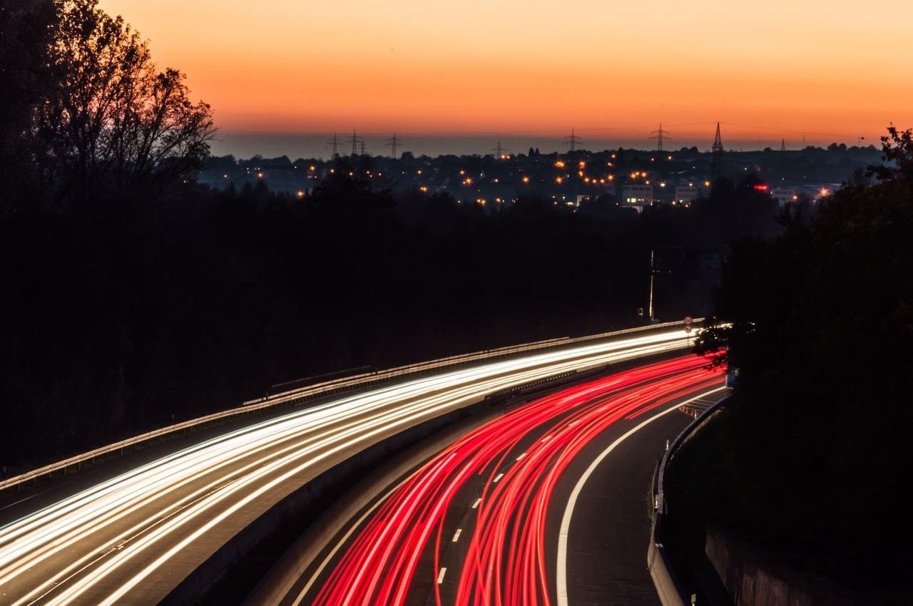 Long-exposure view of a curving highway at dusk, with white and red light trails from vehicles sweeping through the foreground; dark trees line both sides, and a distant town with scattered lights sits under an orange-to-blue sky.
