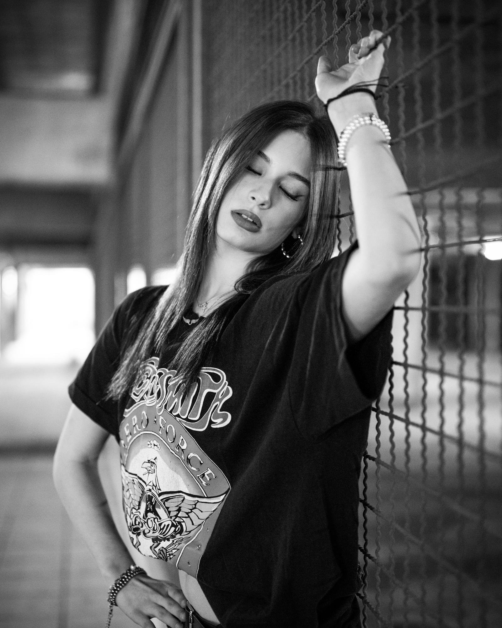 Young woman with long hair wearing a graphic t-shirt, eyes closed, leaning against a chain-link fence. Her left arm is raised, hand grasping the fence. The background is a blurred passageway, giving a sense of depth. The image is in black and white.