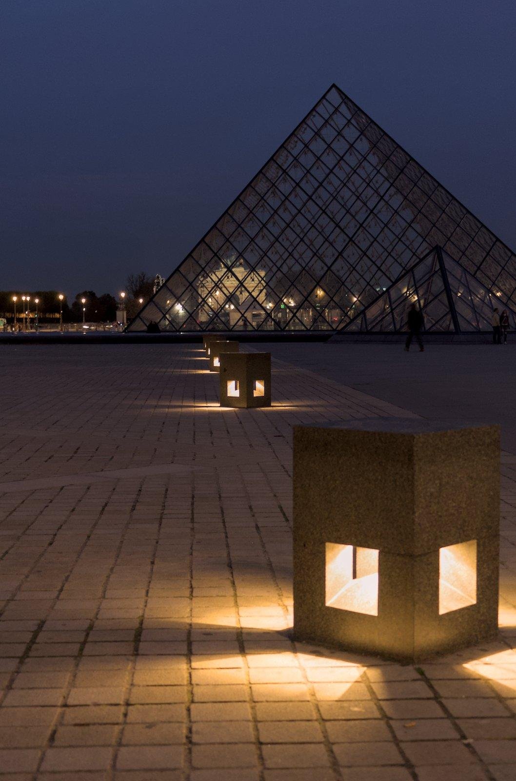 Vertical photo , at night, 3 more or less cubic studs illuminating the ground from their 4 sides, aligned from the lower right corner to the foreground towards the upper left corner, leading to the pyramid of the Louvre occupying half of the background, against a background of midnight blue sky