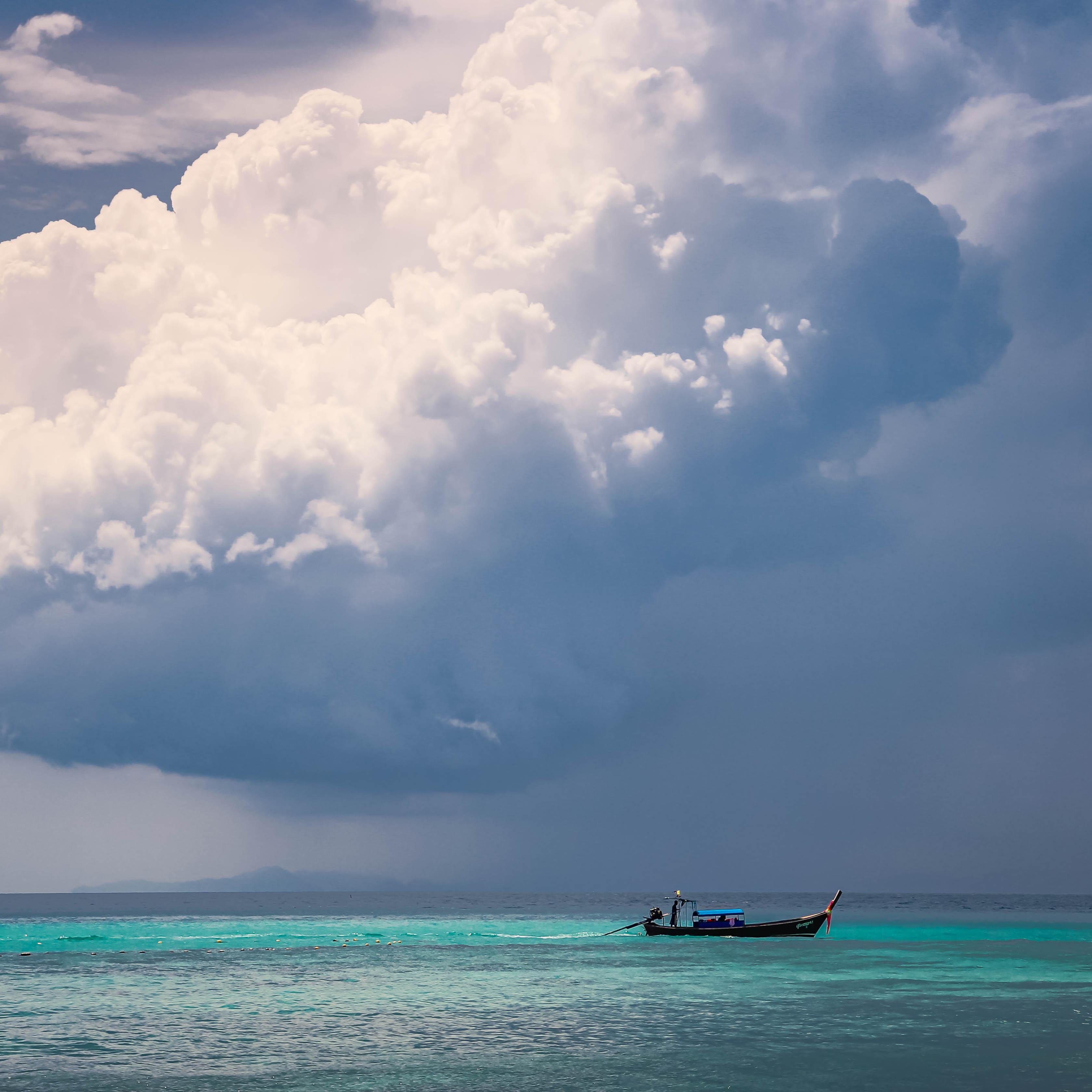 The image shows a scenic view of a large cloud formation over an ocean or sea. There's a small boat on the water, with a person standing in it. The water is a clear turquoise color, creating a contrast with the darker sky and clouds above.