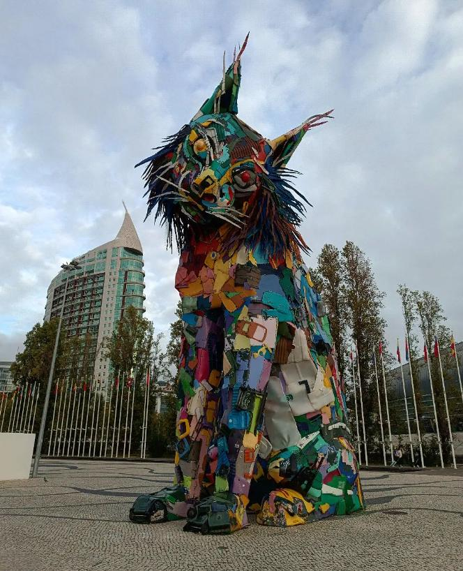 The image features a large sculpture of a lynx made from recycled materials. The colorful sculpture is displayed in an outdoor setting, with a modern building and a row of flags visible in the background. The artwork showcases a creative use of discarded items to create an impressive and vibrant installation.