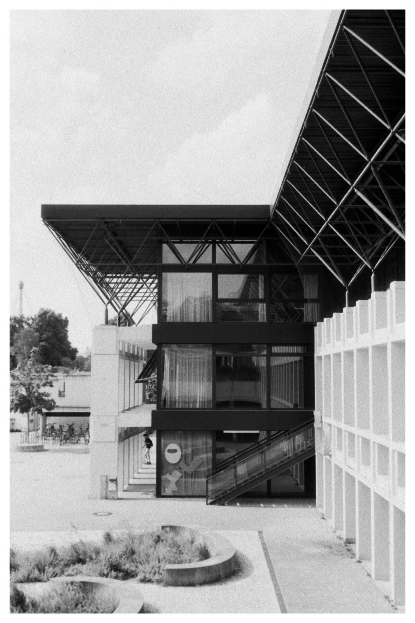 Black-and-white photo of a modern building with a large cantilevered roof supported by exposed steel trusses, glass-walled upper levels, and a diagonal exterior ramp leading to an entrance; concrete planters in the foreground and an open courtyard with trees and bicycles to the left.