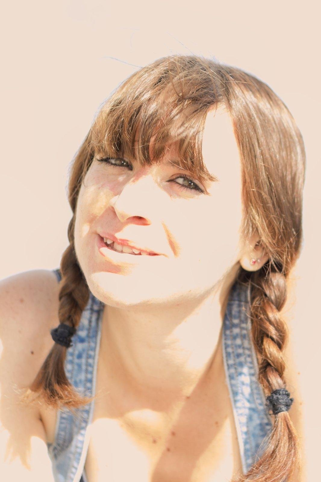 Photo format portrait en couleur, montrant une jeune femme en gros plan, brune, coiffée de deux nattes tombant devant ses épaules, souriante et regardant la caméra. On voit aussi 2 bretelles en denim bleu clair