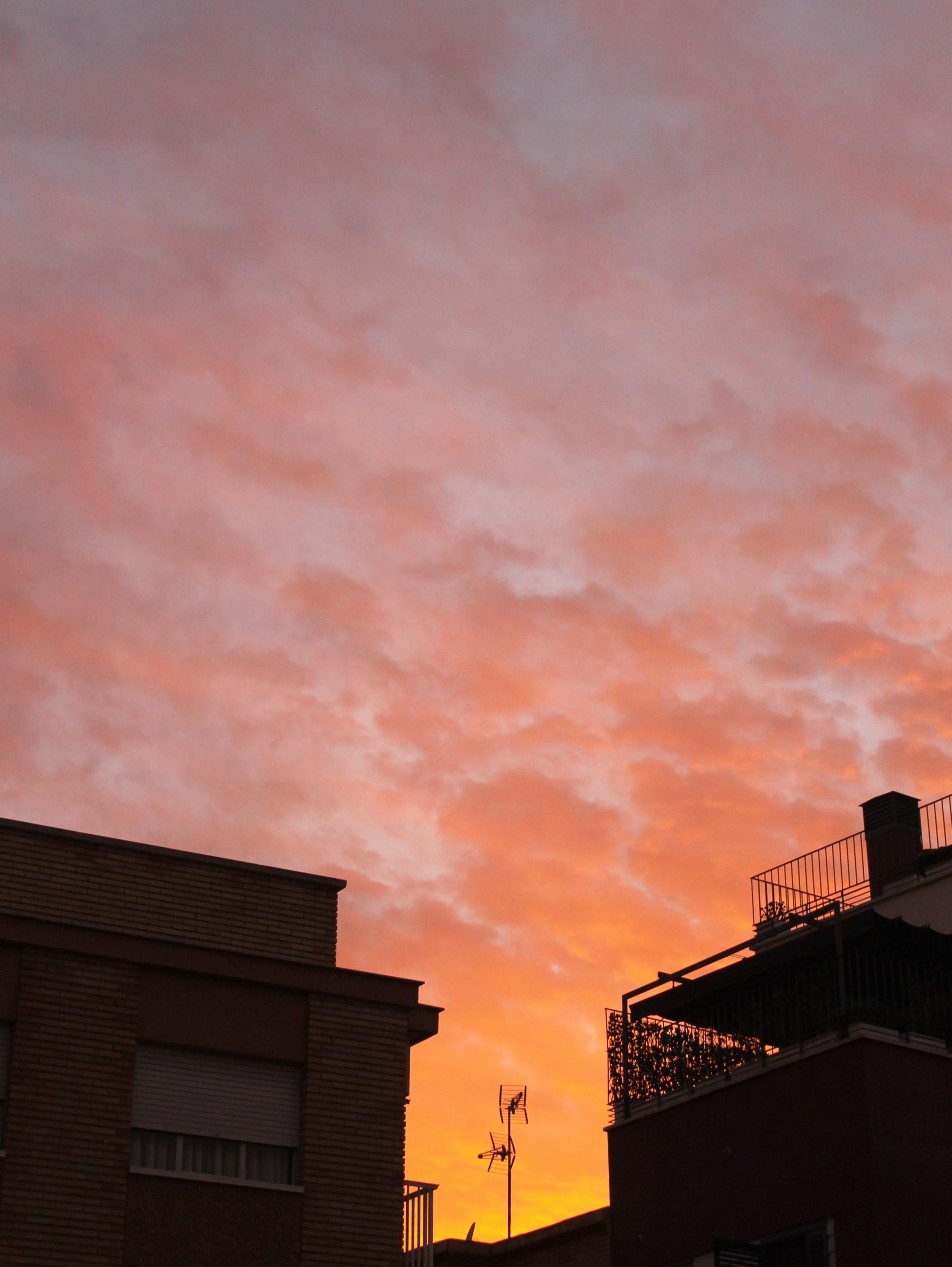 Foto vertical del cielo con nubes anaranjadas y violáceas por una puesta de sol. El sol no se ve, ya se ha escondido entre las siluetas oscuras de unos edificios y antenas de televisión que quedan a contraluz con el cielo