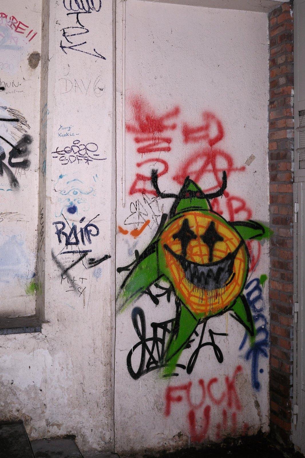 Un graphiti consistant en un disque sommairement griboullé en orange, avec des yeux en étoile et une bouche menaçante. Le disque est agrémenté de triangles verts faisant des membres et un chapeau. Dessous, taggé en rouge «&nbsp;FUCK U&nbsp;»