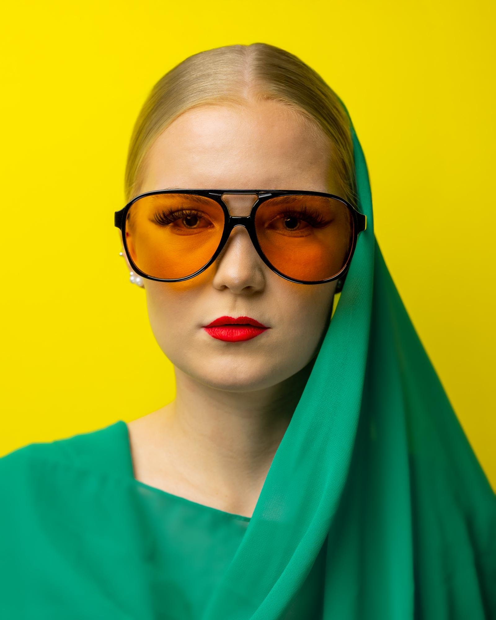 This image shows a person wearing large, amber-tinted sunglasses and a vibrant green garment, which appears to be a wrap or drape. The background is a bright yellow, creating a striking contrast with the colors of the outfit and accessories. The person has bold red lipstick and a neutral expression.