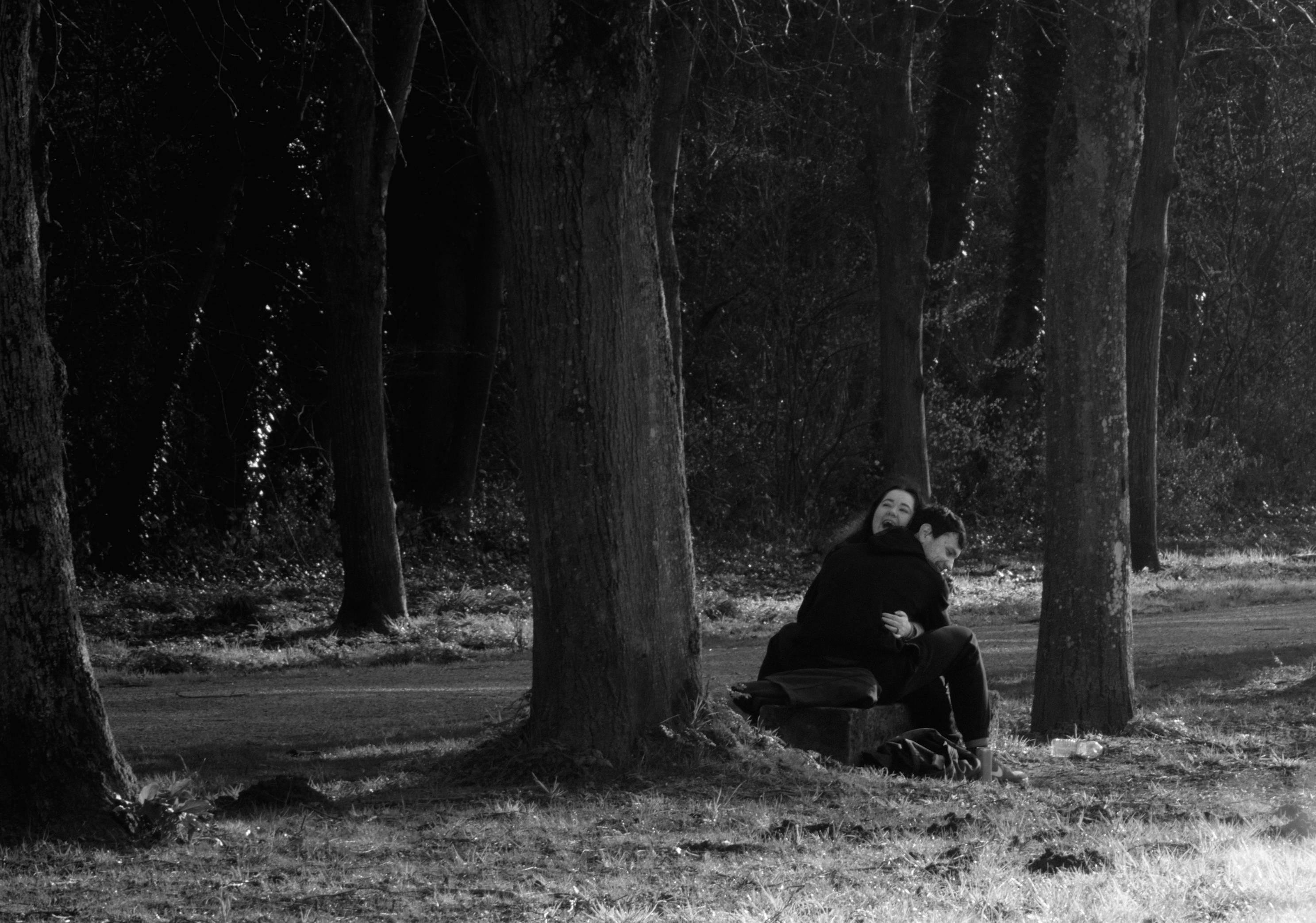 La photographie représente une scène en noir et blanc de deux personnes assises à califourchon sur un banc dans une zone boisée. Elles s'enlacent en riant.