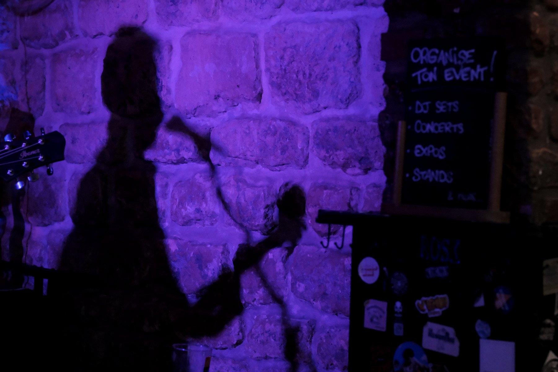 Photo horizontale. Un mur en pierre éclairé par une lumière violette qui projette l’ombre du musicien, le manche de sa guitare et son micro