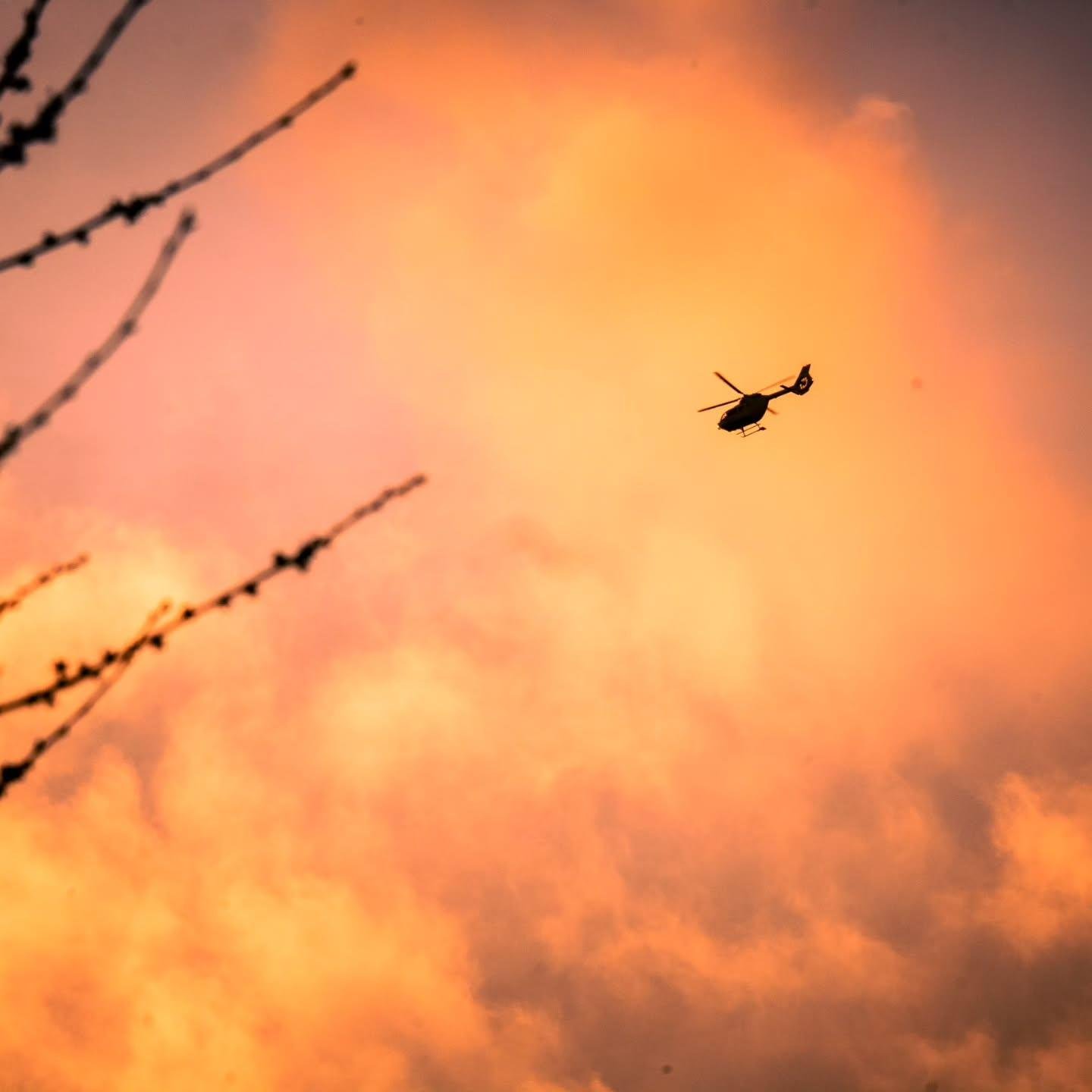 The image shows a helicopter flying against a vibrant orange sky, suggesting sunset or sunrise. There are also some tree branches visible on the left side of the image.
