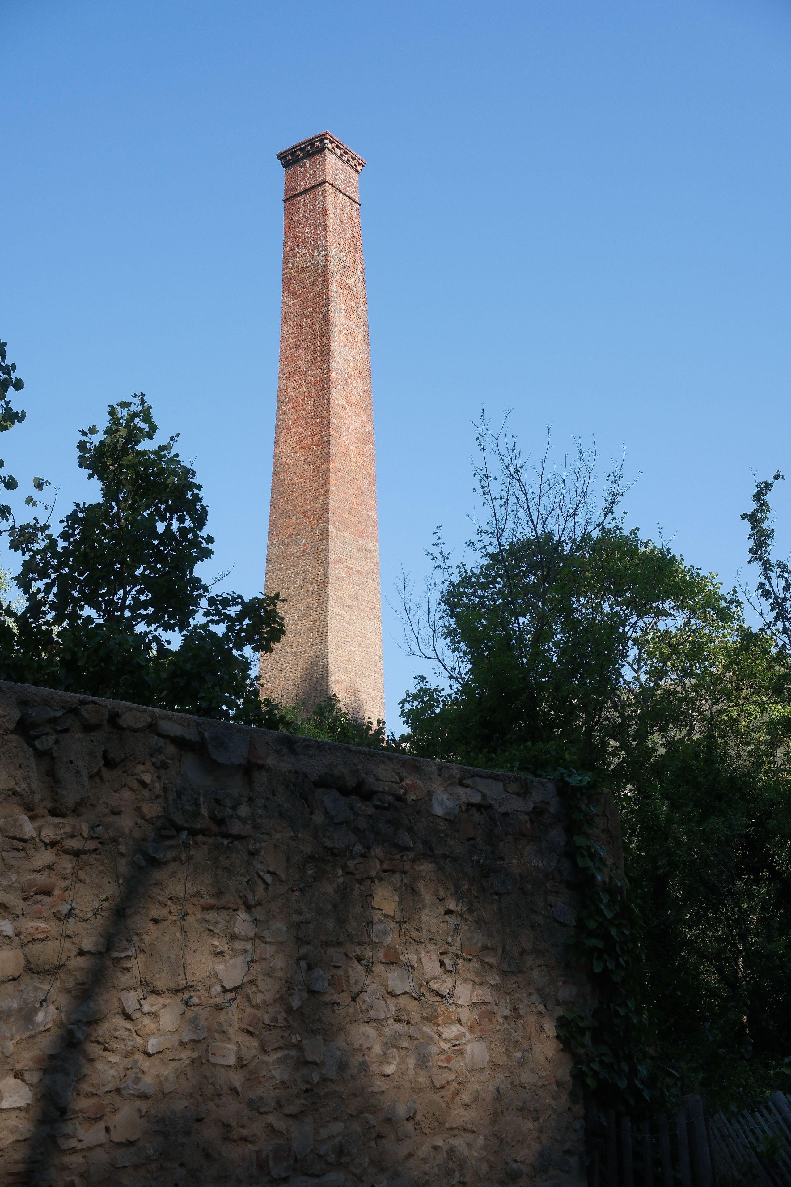 Portrait format photo. The foreground is in the shadows and we see an old light brown stone wall. Above it, some vegetation and behind it, a red-ish square chimney with a clear blue sky all around