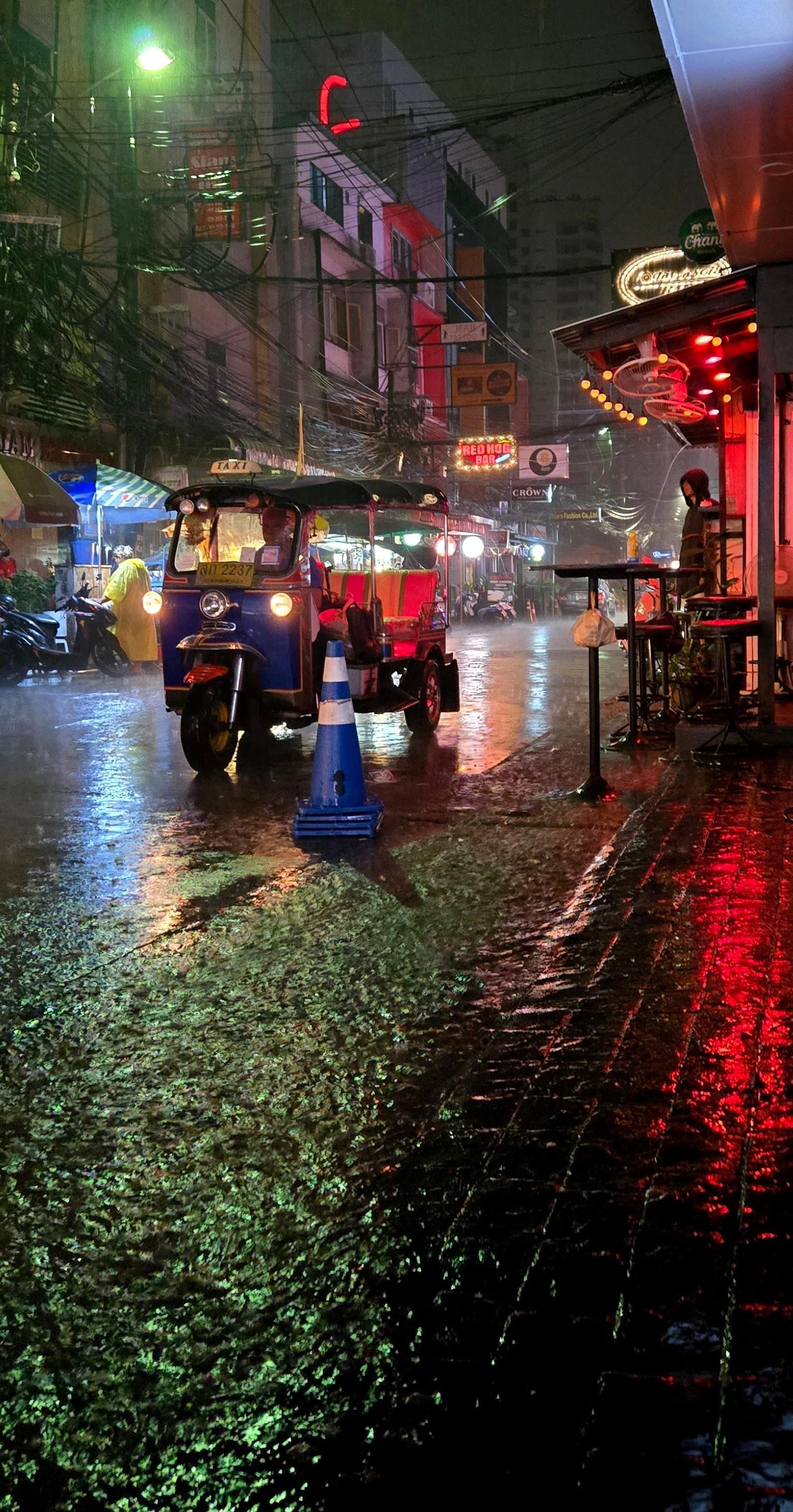 Rainy night street scene with a blue tuk-tuk taxi stopped near a blue-and-white traffic cone, wet pavement reflecting red and green neon lights, tangled overhead power lines, and bar signs and storefronts along the right-side sidewalk.