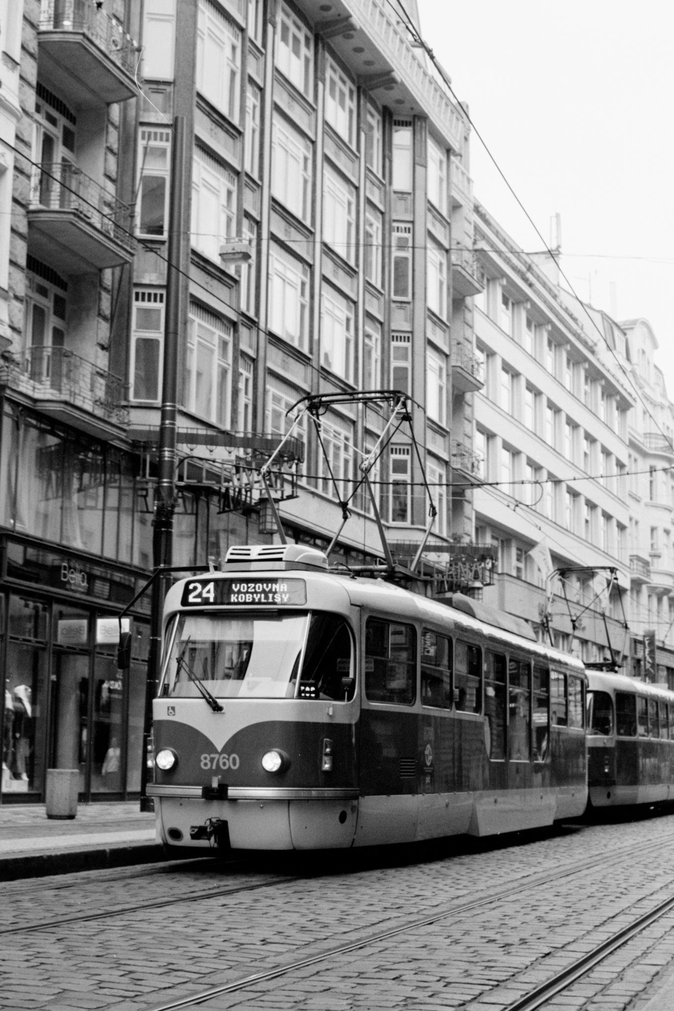 Black-and-white street scene with a tram on cobblestone tracks, overhead power lines above it, and tall multi-story buildings with balconies lining the street; the front display reads “24” and “VOZOVNA KOBYLISY,” with car number “8760” visible.