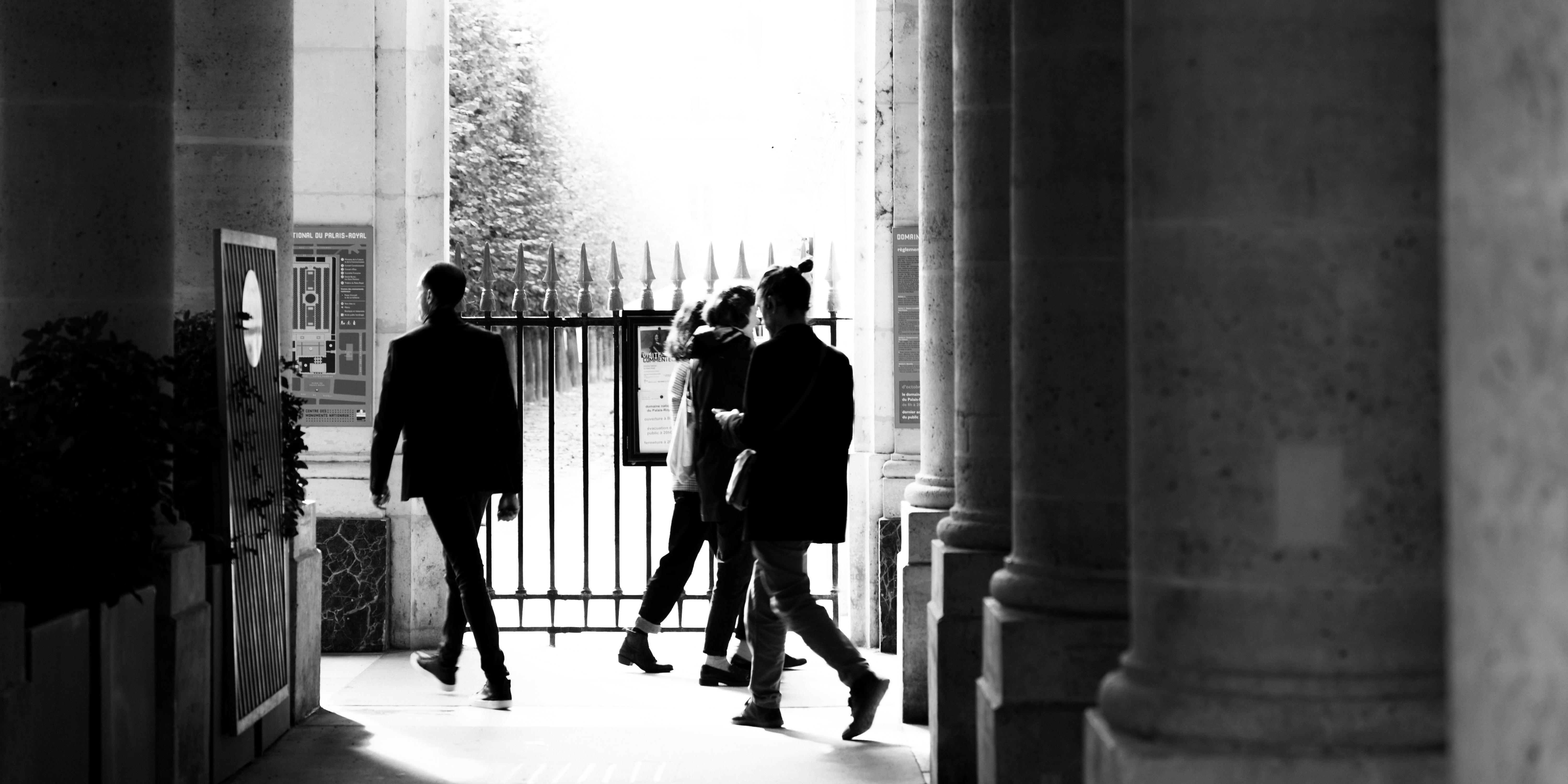 Three people walking through a colonnade with tall stone columns on either side, approaching a metal gate with spikes. The scene is backlit, creating silhouettes of the individuals. Potted plants are visible on the left, and light filters through trees in the background.