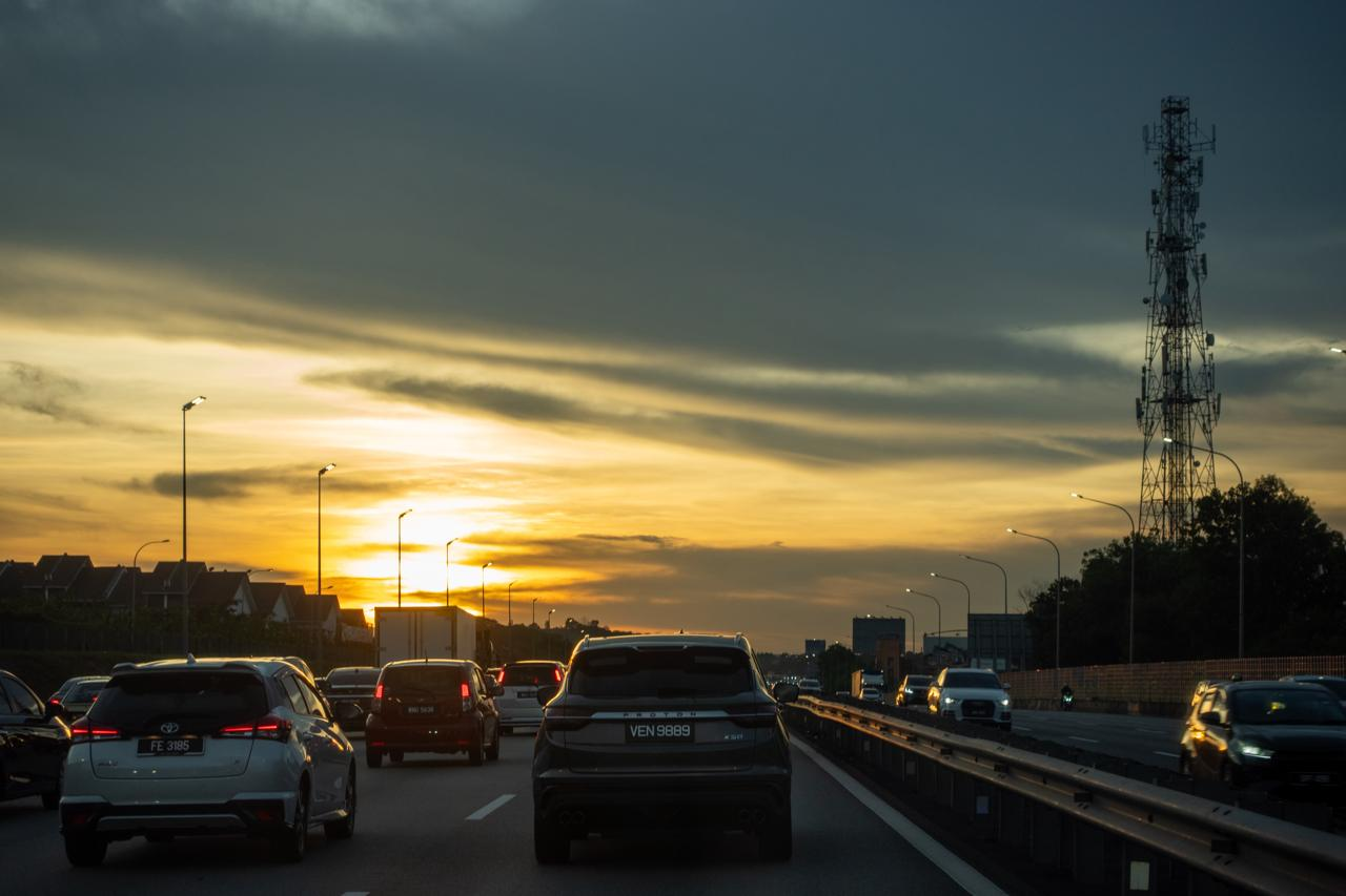 Traffic moving on a highway during sunset, with cars in different lanes. The sky features a mix of orange and dark clouds. Streetlights line the road, and a tall telecommunications tower is visible to the right. Trees and distant buildings are silhouetted against the colorful sky.