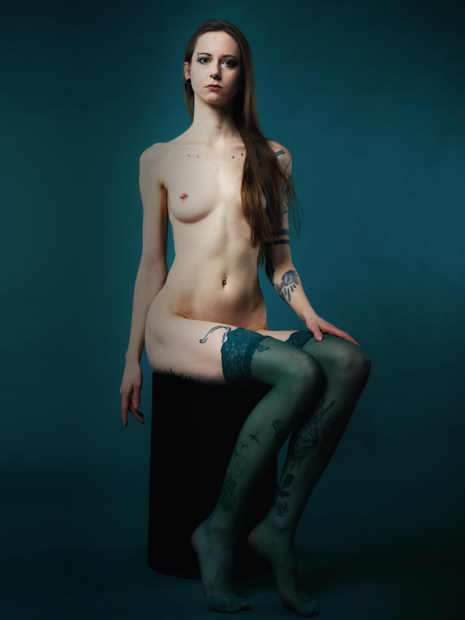 Young tattooed and pierced naked woman, dressing only green stockings, full portrait sitting on the chair.