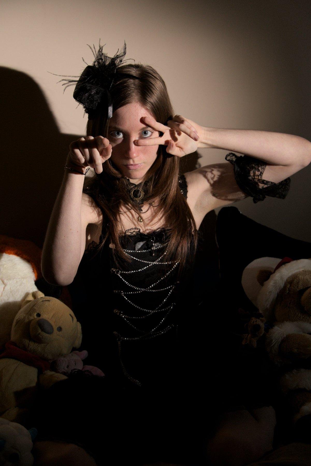 Photo format portrait montrant une jeune femme rousse (mais l’éclairage rends brune) habillée avec une barrette ornée d’un petit chapeau noir avec plein de froufrou, un collier type chocker avec un fermoir en forme de cercle doré entourant une pierre noire, un haut faisant idole japonaise, noir avec des chaîne se croisant sur le torse, et un brassard en dentelle sur le bras gauche. Elle est assise sur un lit et entourée d’ours en peluche, sa main gauche forme un V horizontal autour de son œil gauche et la main droite pointe la caméra. Elle regarde droit vers la caméra, la tête légèrement penchée vers le bas. La scène est éclairée par un fort spot projetant l’ombre du modèle contre le mur beige derrière