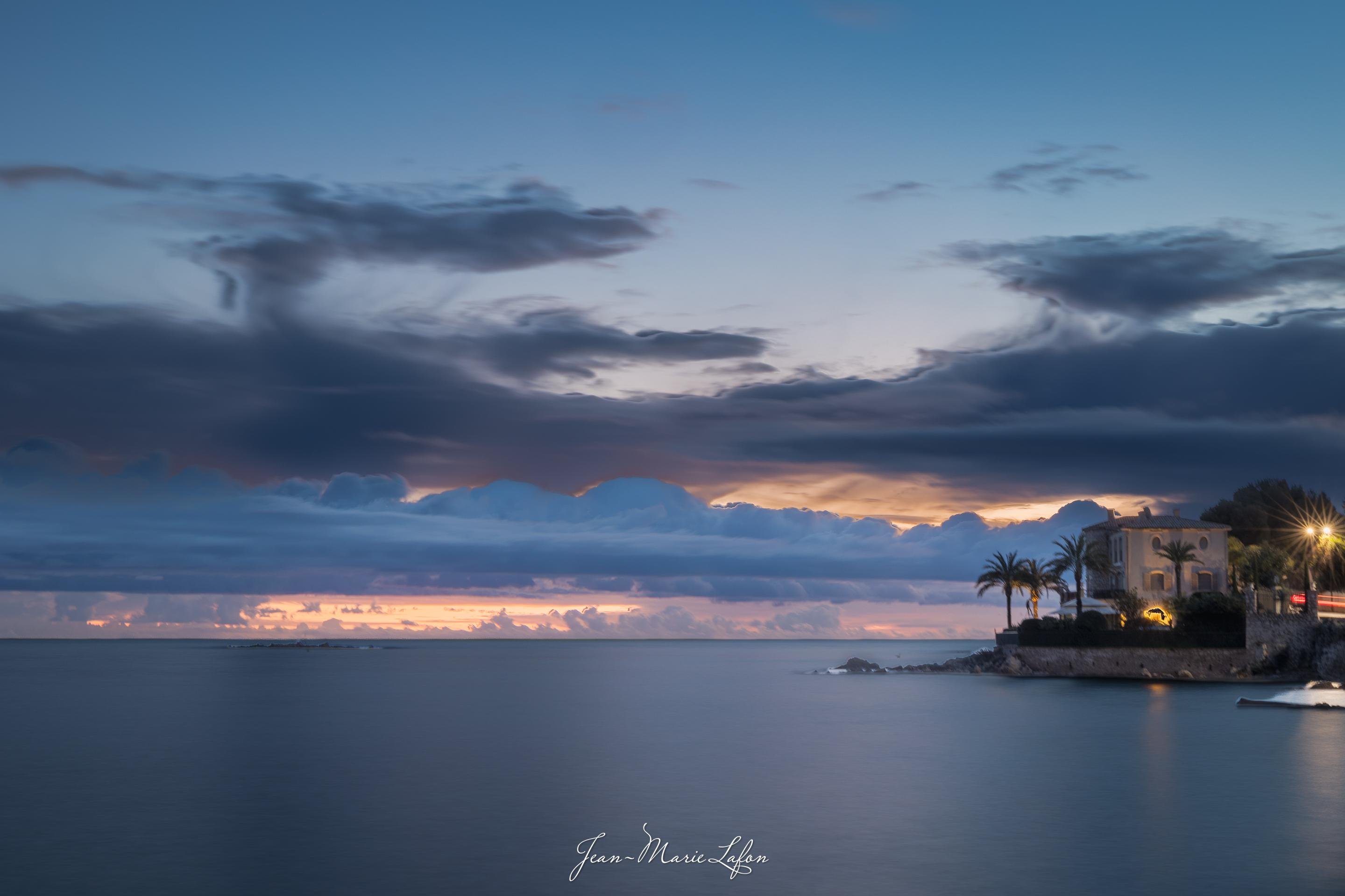 Coastal seascape at dusk with calm water in the foreground, layered dark clouds across a blue-and-orange horizon, and a small rocky point on the right with a lit house, palm trees, and a streetlamp.