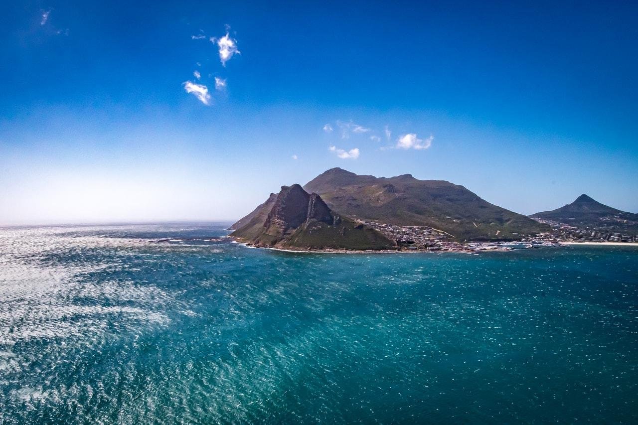 The image shows a coastal landscape with clear blue skies and expansive ocean waters. There is a prominent, rugged mountain or hill extending along the coastline, and a town at its base. The area appears to be a scenic, possibly popular destination with a mix of natural beauty and human habitation.
