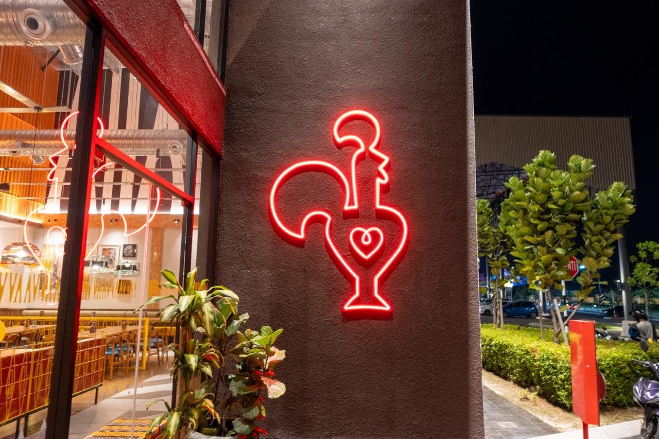 Neon red rooster sign with a heart in the center on a dark wall exterior; indoor view through large window showing tables and chairs. Green plants are visible next to the wall, and trees line a parking lot in the background during nighttime.