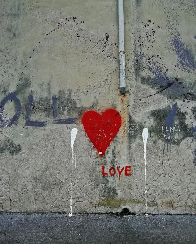 The image depicts a wall with graffiti. There is a large red heart with the word "LOVE" written below it. The wall also has other markings and spray paint splatters, including some drips of white paint.