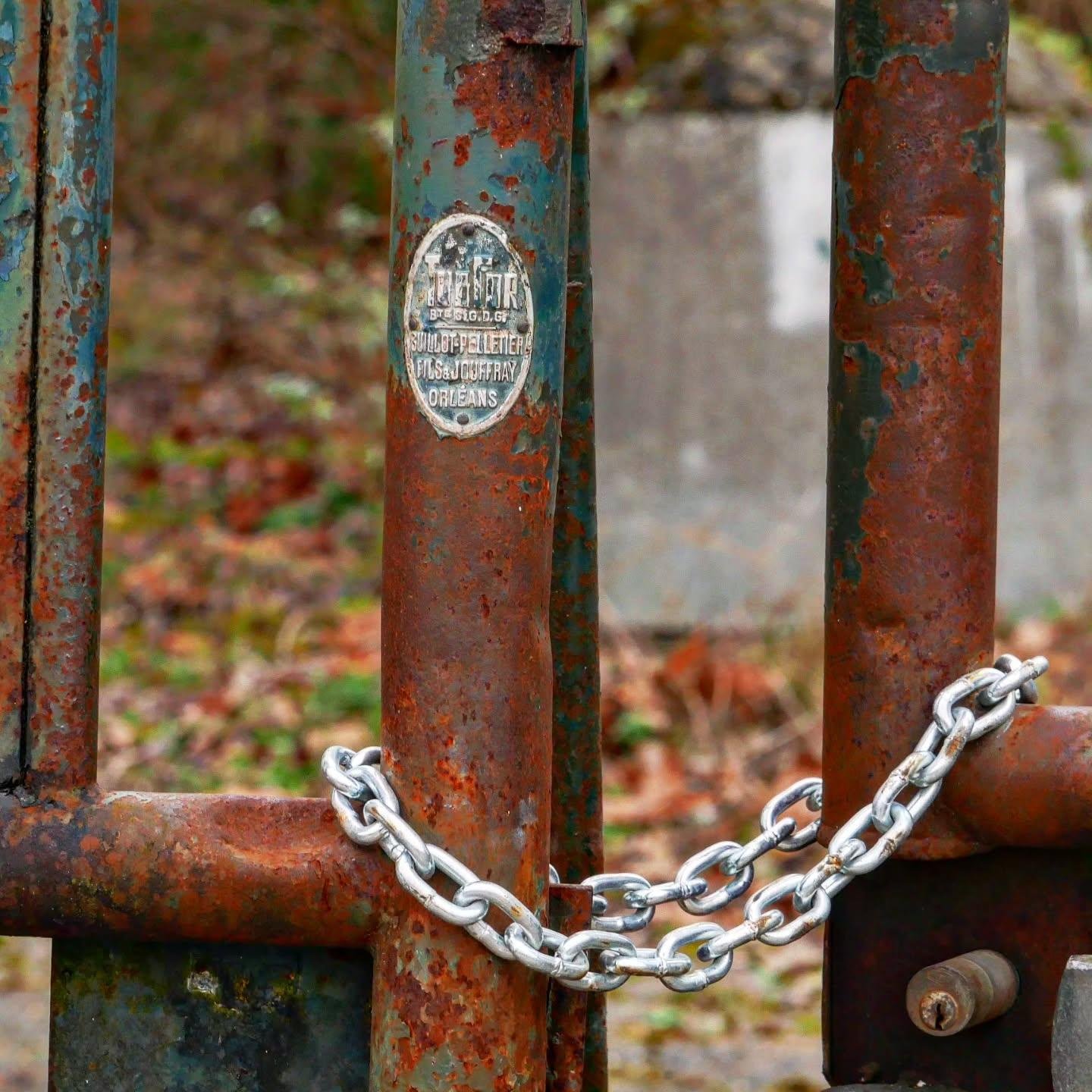 The image shows a close-up of a rusty metal gate secured with a shiny chain. The chain is wrapped around two bars of the gate, and there is a sign on the gate with some text, though the rust makes it difficult to read. The background is blurred, showing some foliage and possibly a concrete structure.