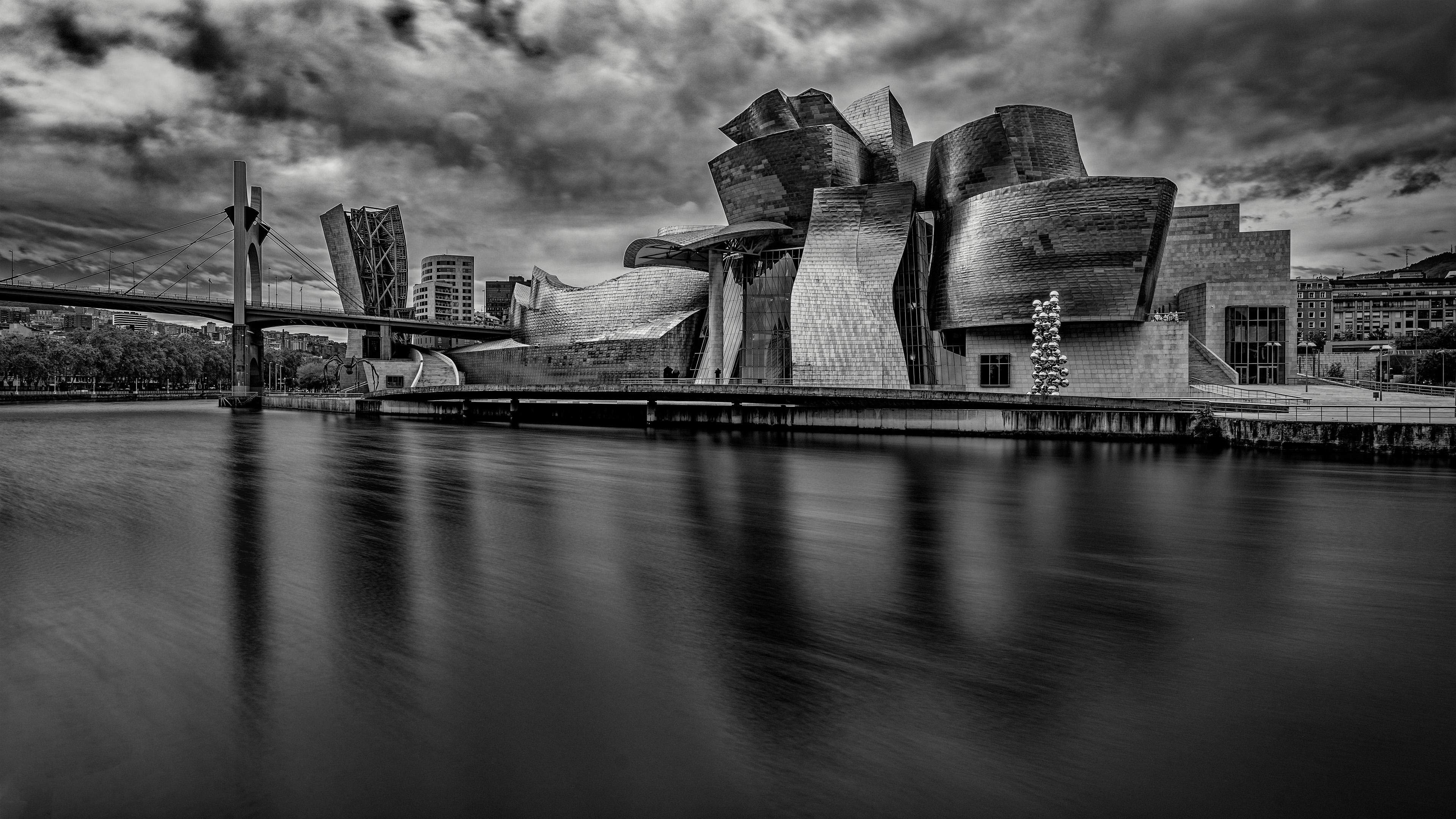 Modern architectural building with a striking design, featuring metallic, curved shapes and reflective surfaces. Positioned alongside a calm river with a smooth water effect. A bridge extends across the left side, while cloudy skies form the backdrop. An arrangement of circular art pieces stands near the structure.