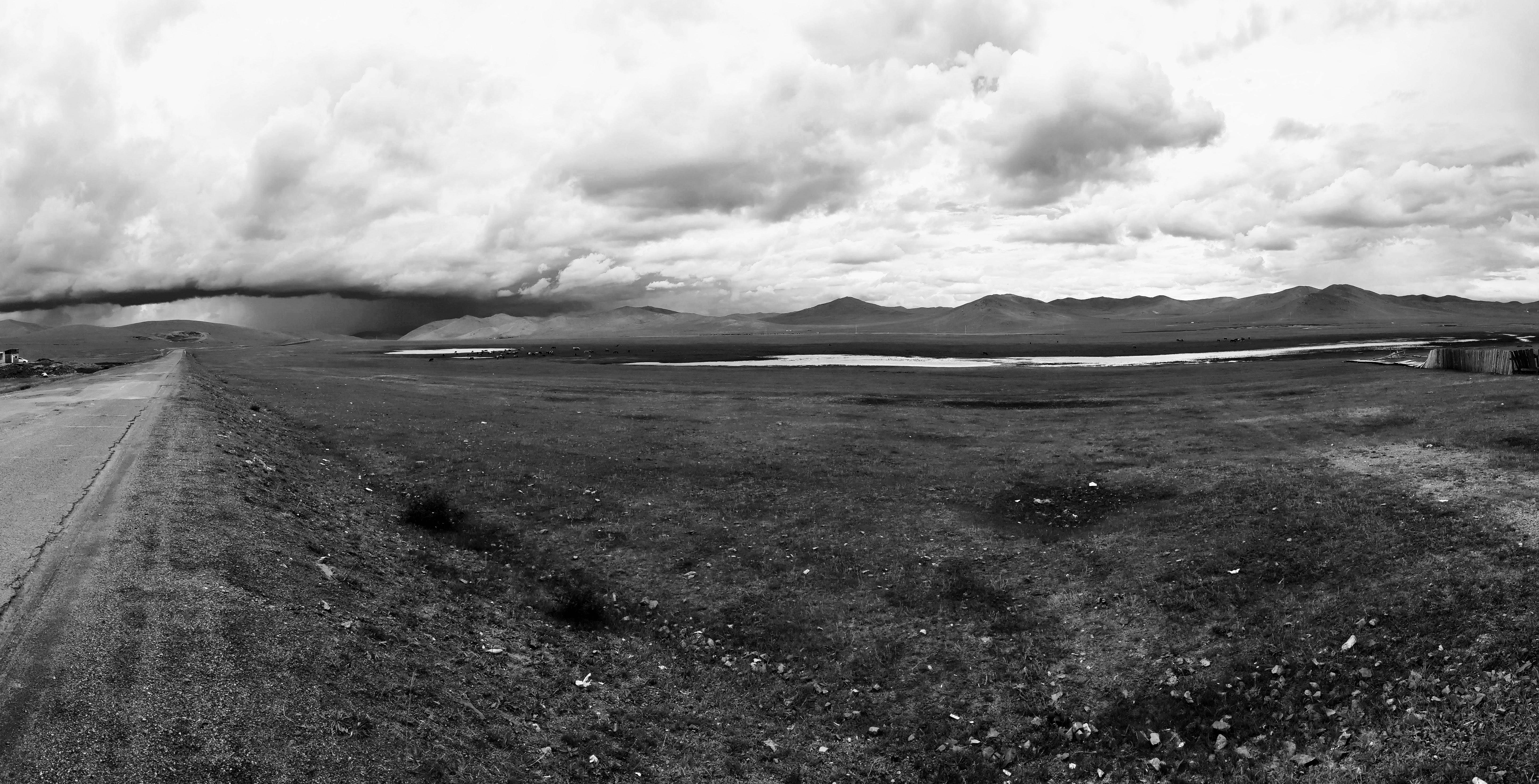 Black-and-white panoramic landscape with a straight road along the left edge, open grassy plain in the foreground, a winding river or lake across the middle, distant low mountains on the horizon, and thick storm clouds with a dark rain band on the left.