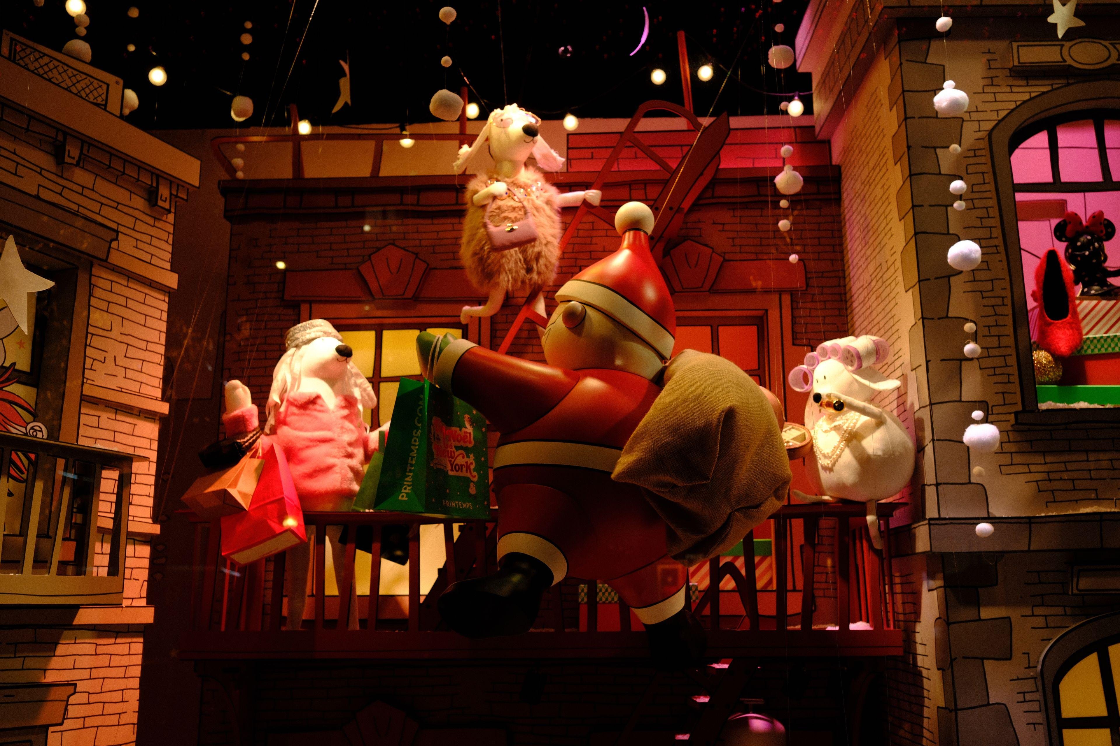 Photo horizontale. Décorations de Noël d’une vitrine. Un père Noël entrain de livrer des cadeaux au balcon d’un immeuble où des chiennes humanoïdes les réceptionnent