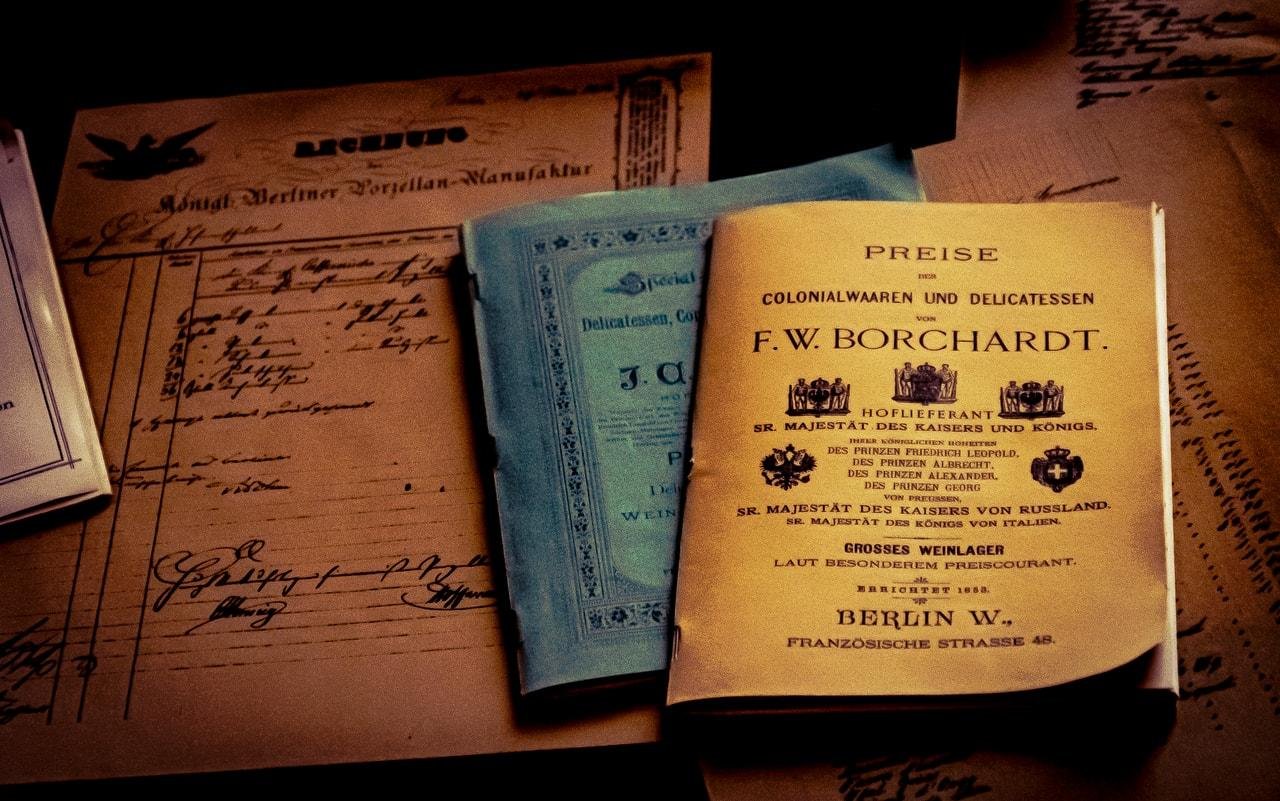Stack of aged paper documents and booklets on a dark surface, featuring handwritten cursive letters and a yellow German price list titled “PREISE … F. W. BORCHARDT” with decorative emblems, partially overlapping a blue booklet.