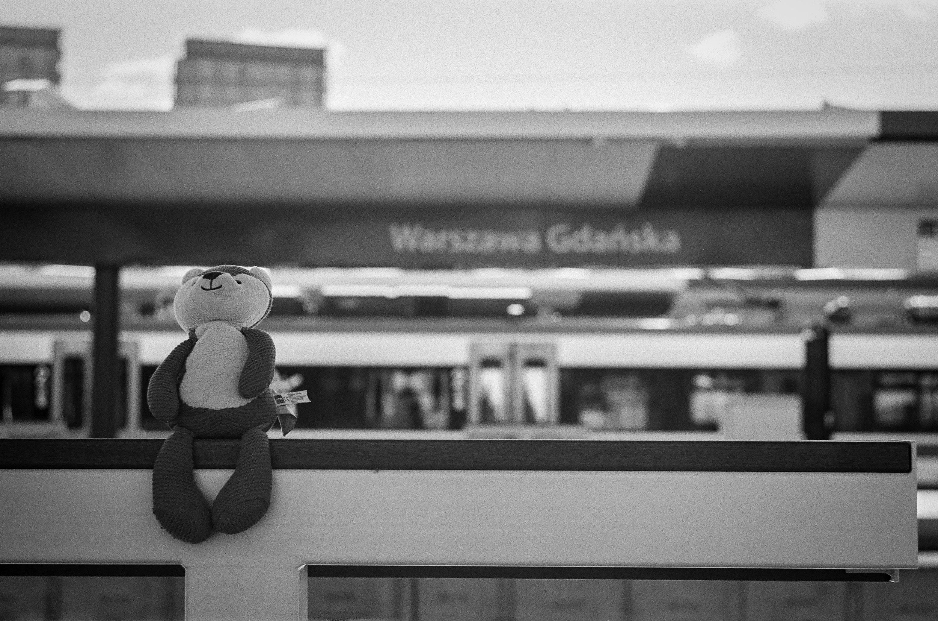 B&W film photo of a fox plushie sat on a leaning "bench" in front of Warszawa Gdańska train station sign