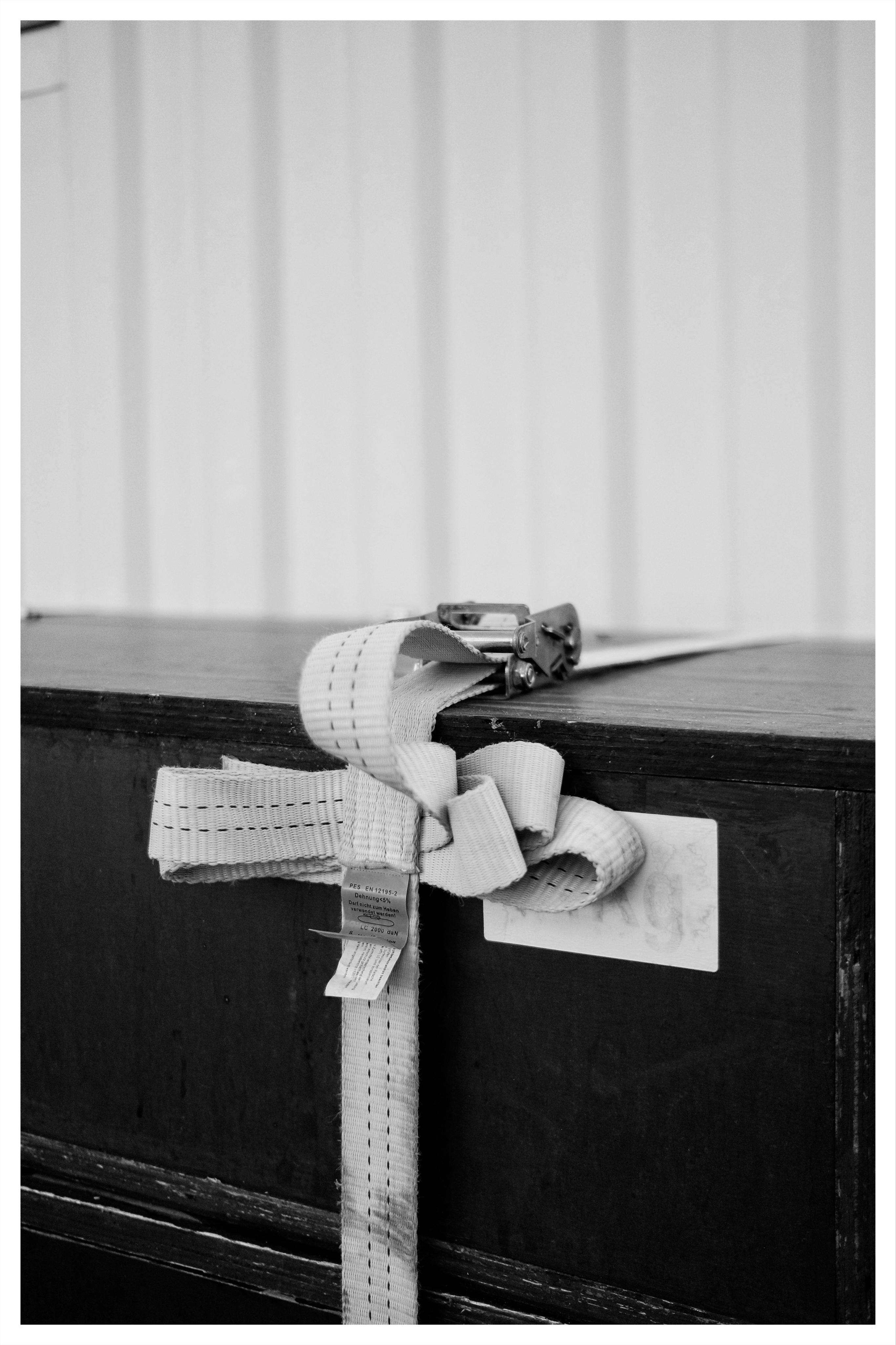 White woven tie-down strap with a metal ratchet buckle looped and knotted around the corner of a dark wooden cabinet, with small attached tags, against a light curtain background.