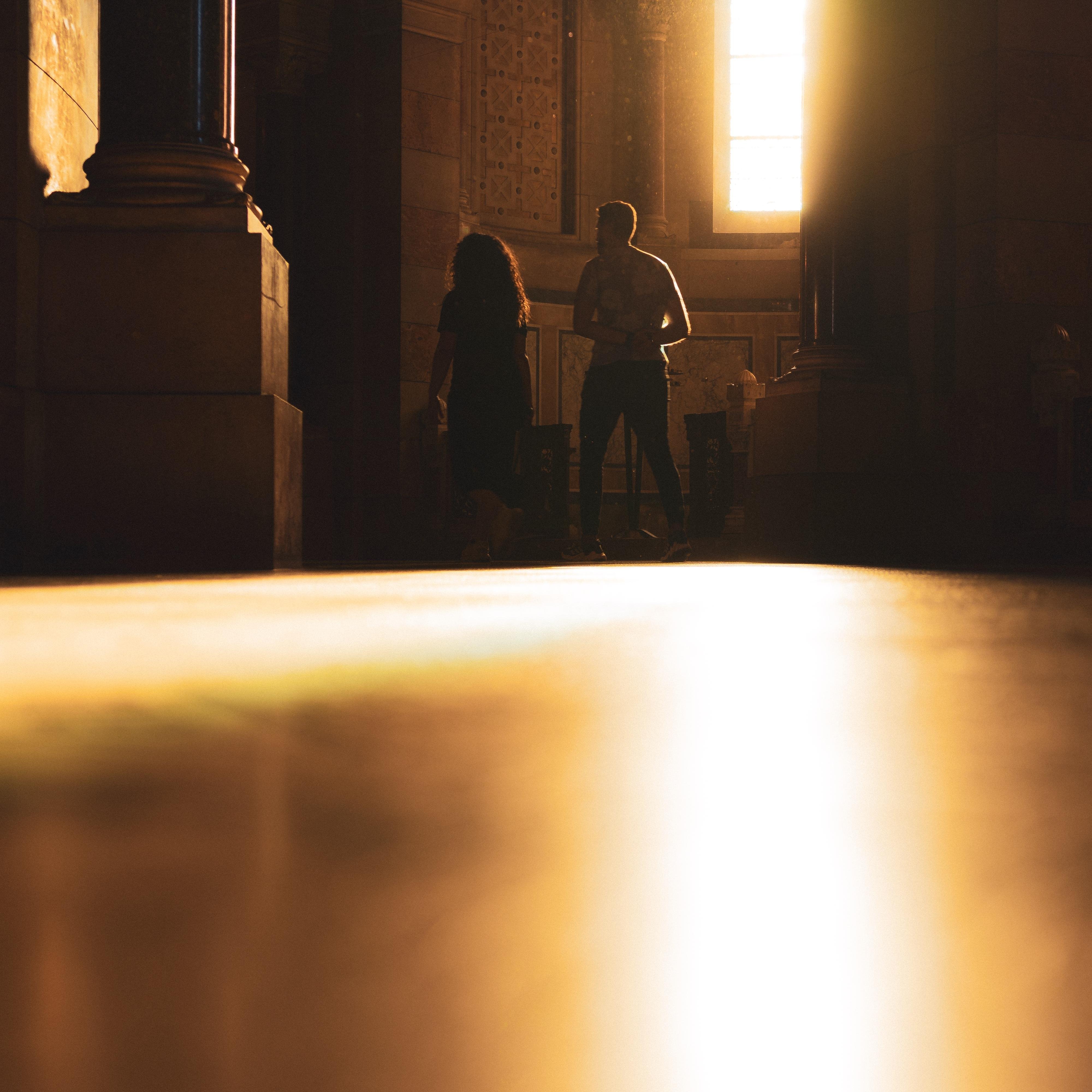 The image shows the interior of a dimly lit room with two people silhouetted against bright light coming from a window. The light creates a dramatic effect, casting shadows and illuminating parts of the room. The foreground appears out of focus, emphasizing the light and figures in the background.