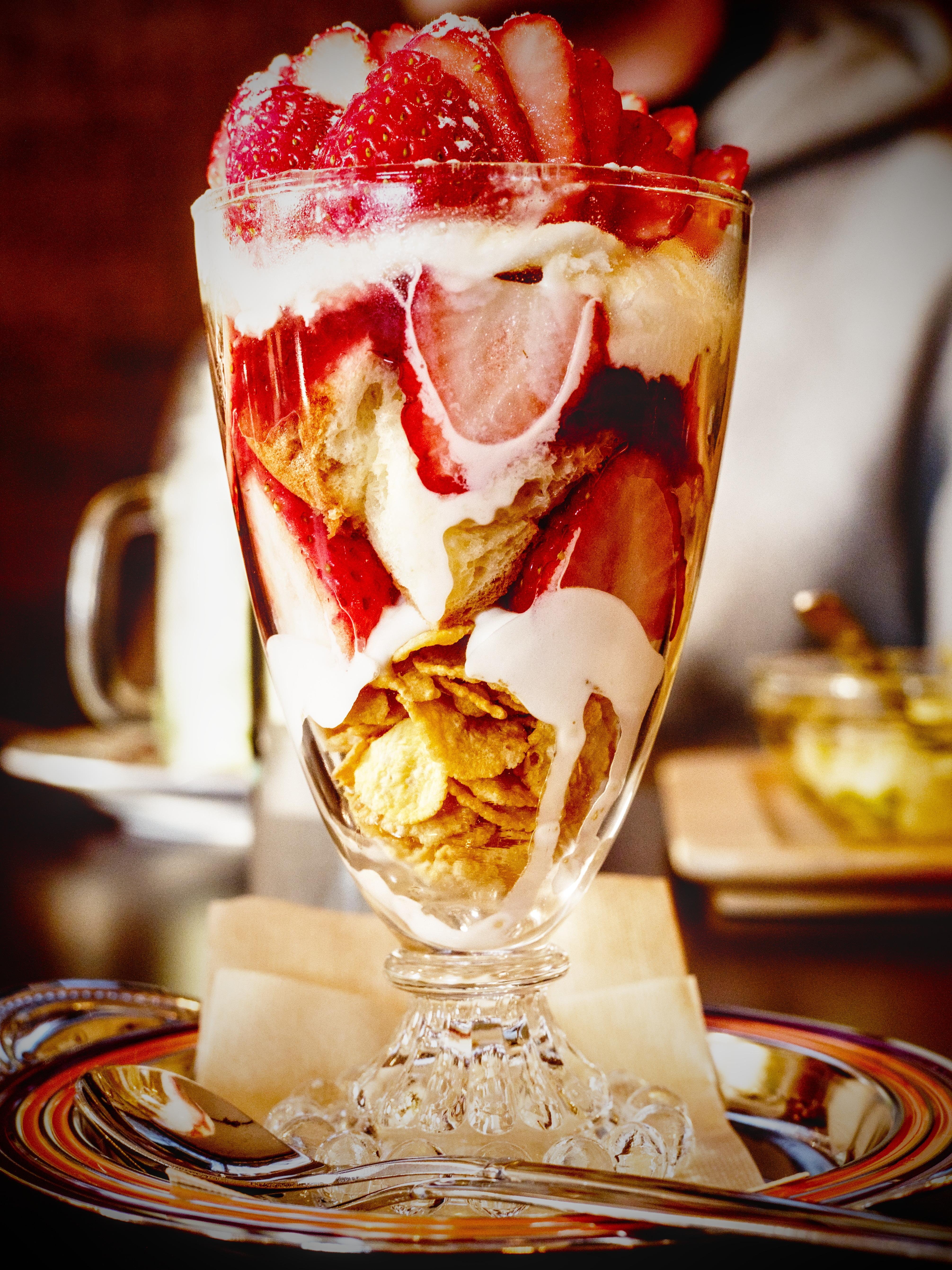 A cup of fresh strawberries with yogurt and corn flakes. The glass holding the contents is taken from the side, exactly in the middle of the frame, and takes up almost the entire vertical space, with the exception of the spoon lying on the plate underneath.