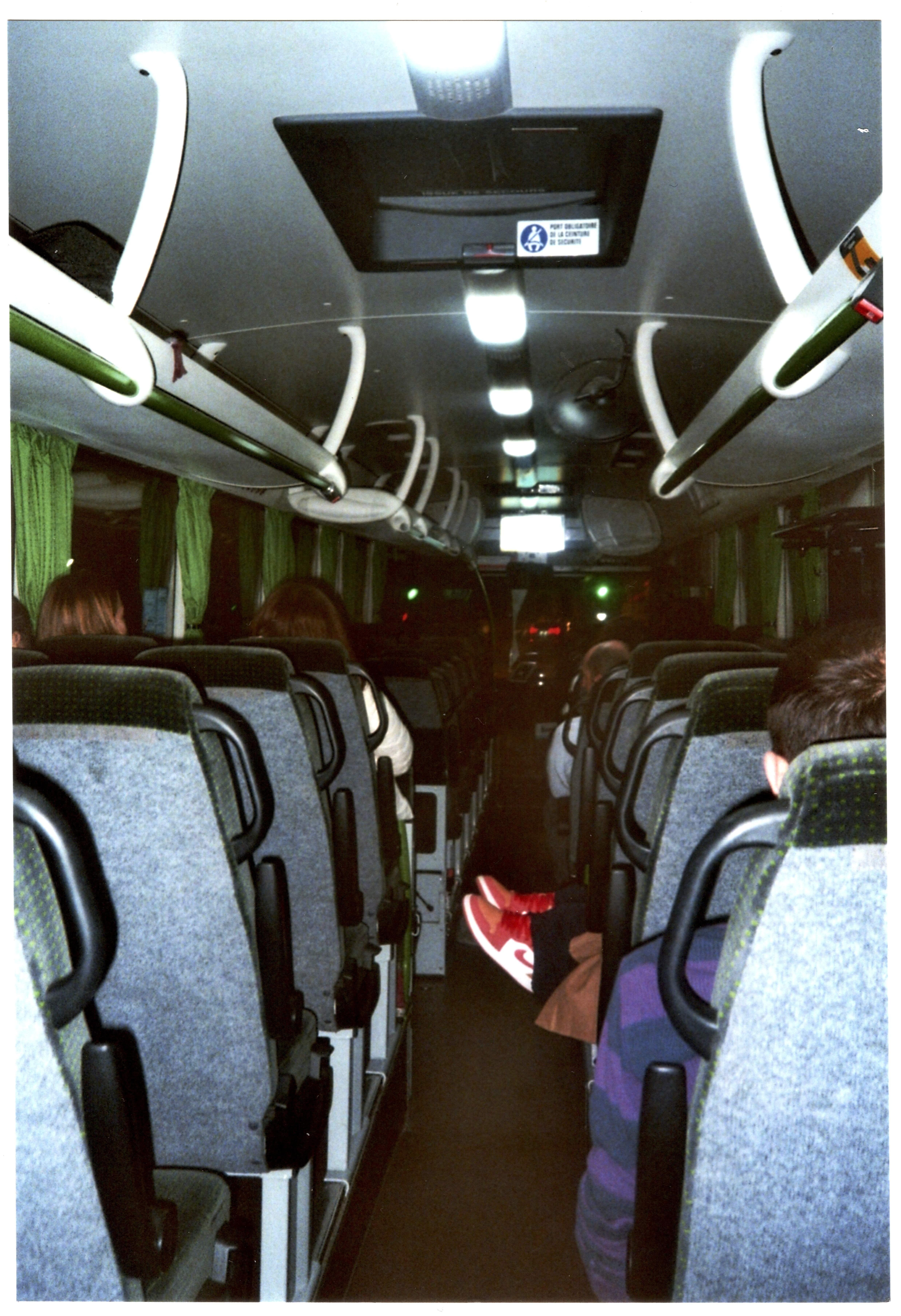 Cette image montre la photographie de l'intérieur d'un bus pris depuis le dernier siège, vers l'avant. Le plafond est équipé de mains courantes et l'éclairage est contrasté. Une paire de chaussures rouges Nike dépasse dans l'allée, venant, on le devine, d'un passager allongé sur des sièges à droites.